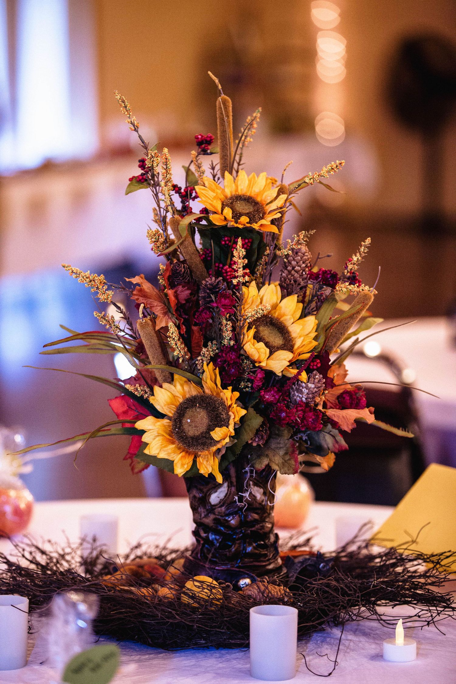 Autumn wedding centerpiece with sunflowers, burgundy blooms, wheat, and candles arranged in a rustic vase on a white tablecloth.