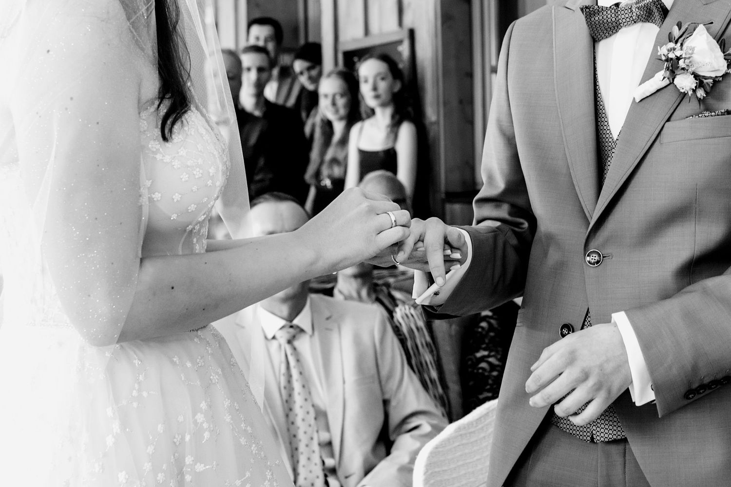 Black and white photo of bride placing wedding ring on groom's finger during ceremony with guests watching.