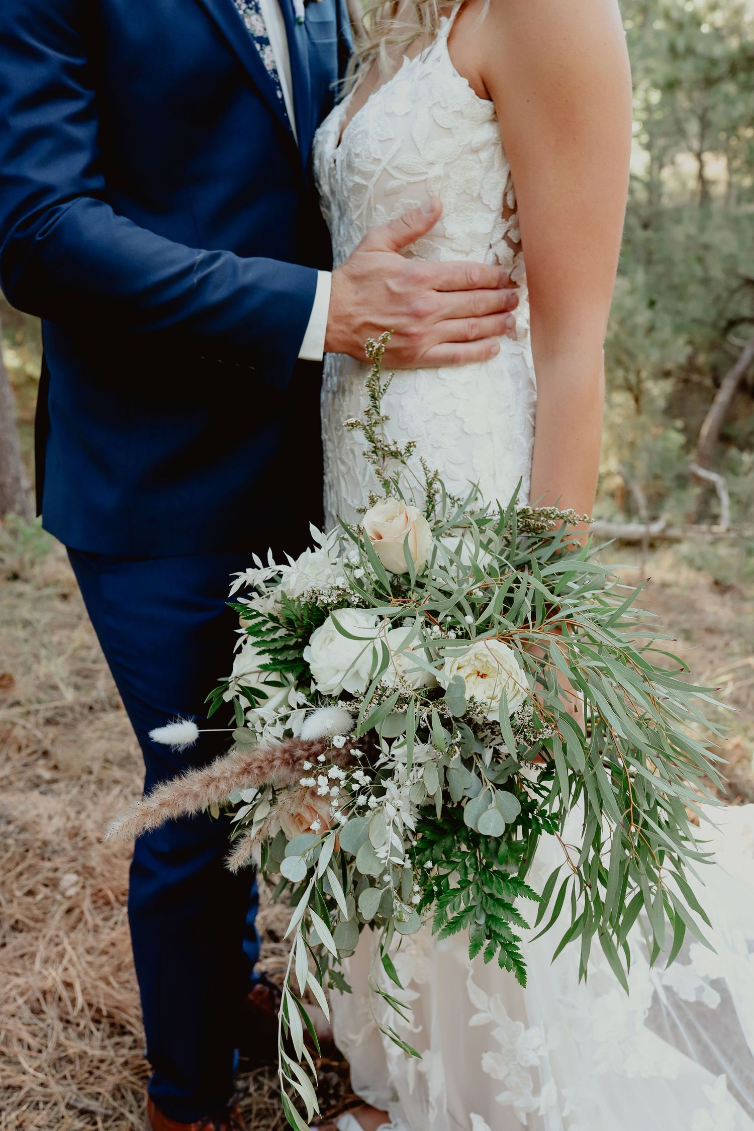 Bride in lace gown holds lush greenery bouquet with white roses as groom in navy suit embraces her waist outdoors.
