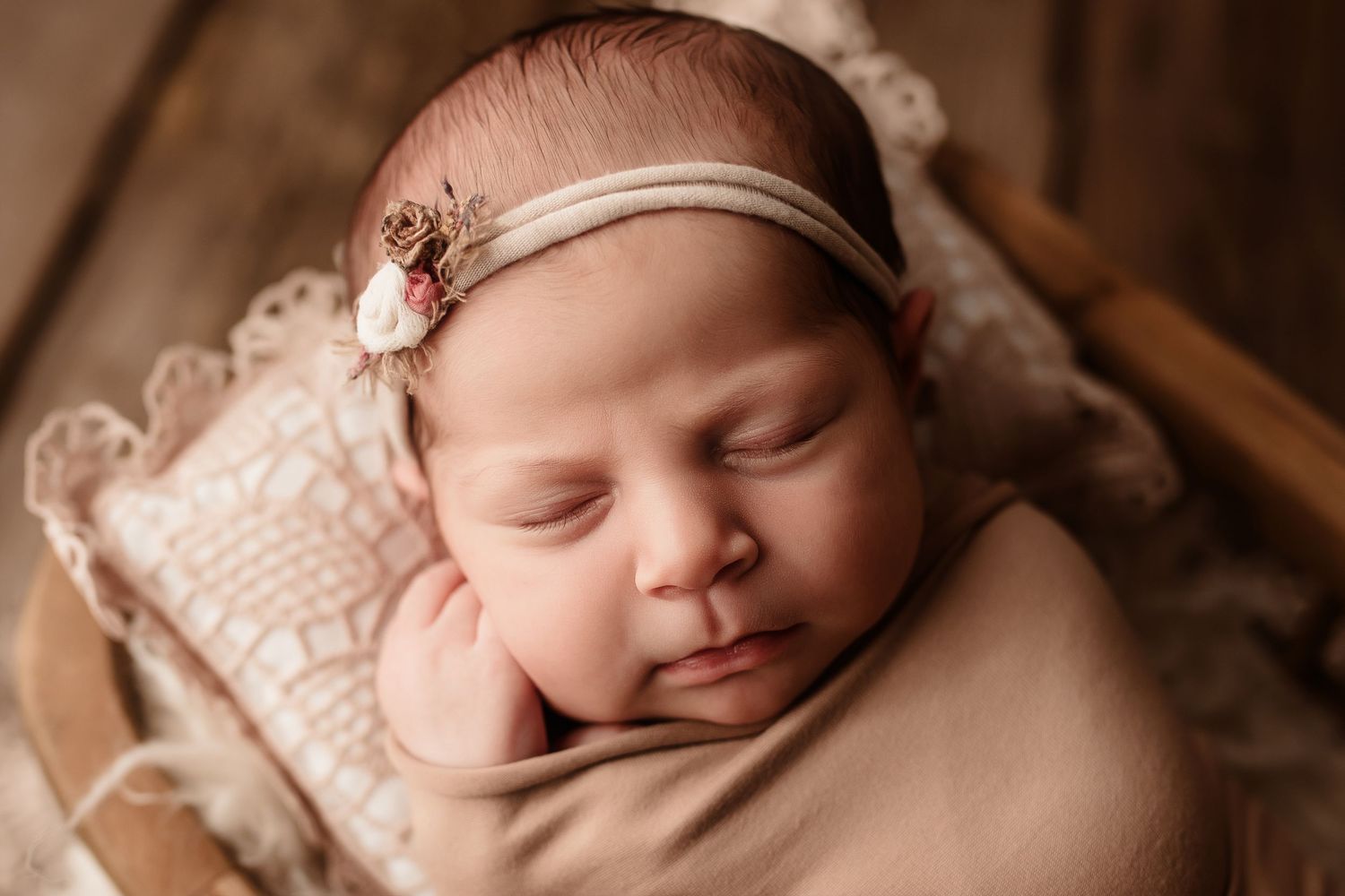 Sleeping newborn baby girl wearing a floral headband nestled in a wooden bowl with lace.
