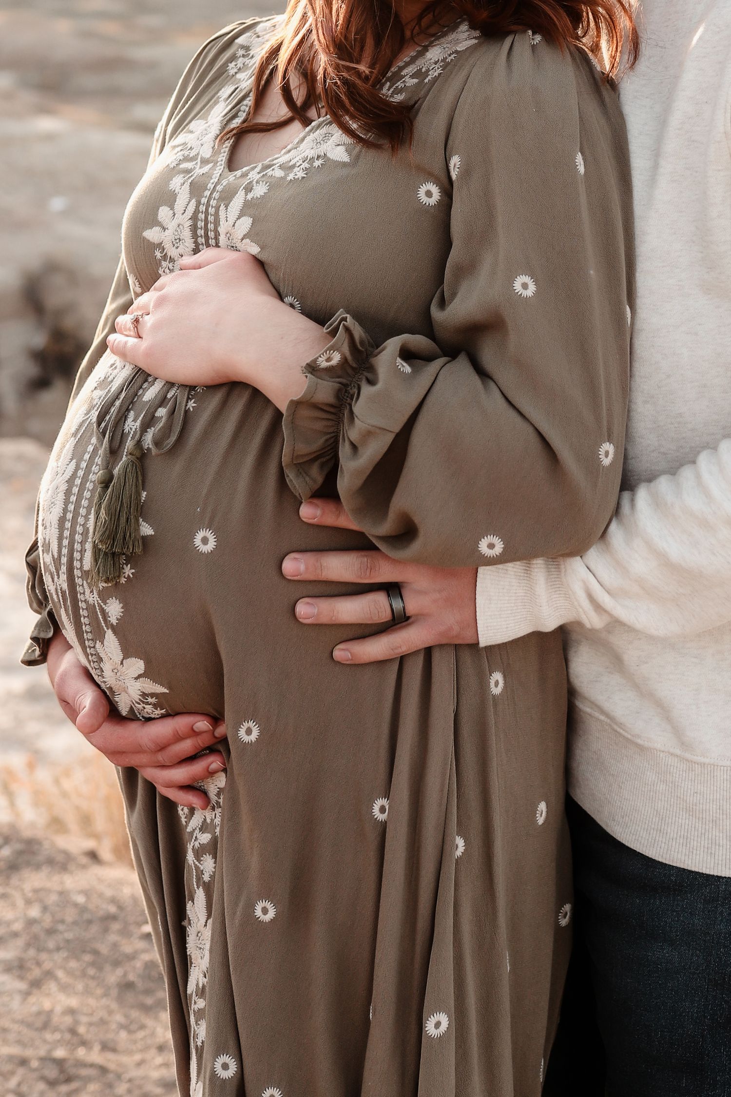 Pregnant woman in embroidered olive dress with partner embracing her baby bump from behind.