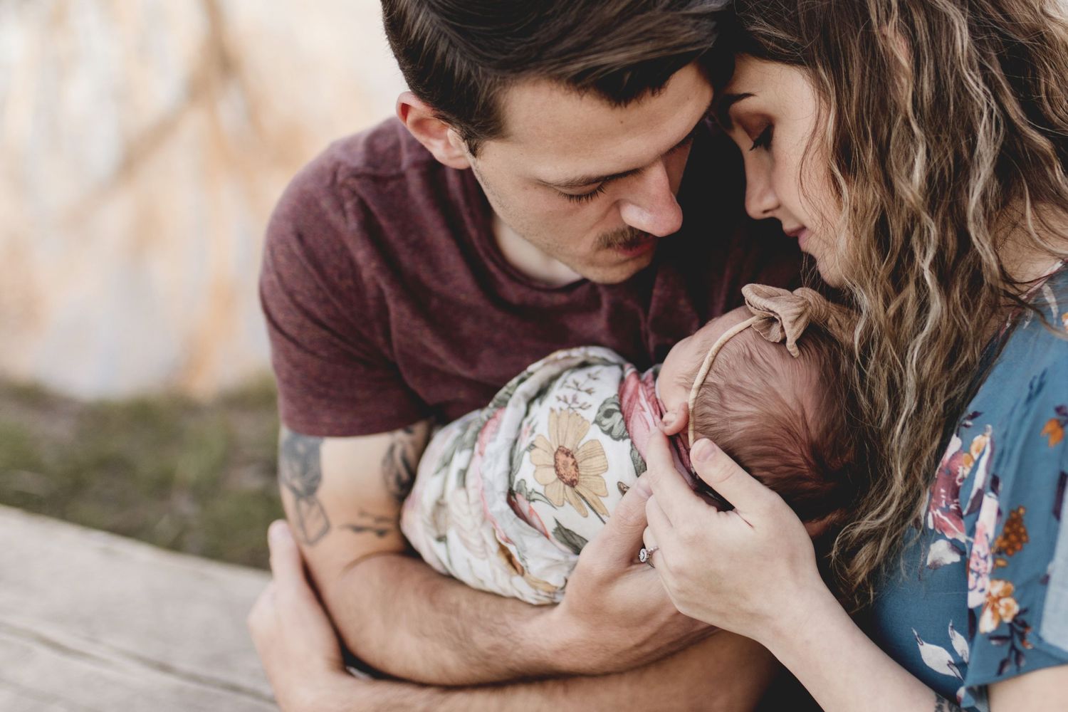 Young couple with tattoos lovingly gazing at their swaddled newborn outdoors.