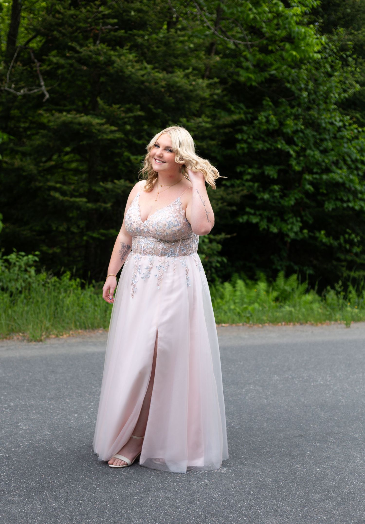Young woman in elegant pink beaded gown with slit poses on outdoor path surrounded by lush green trees.