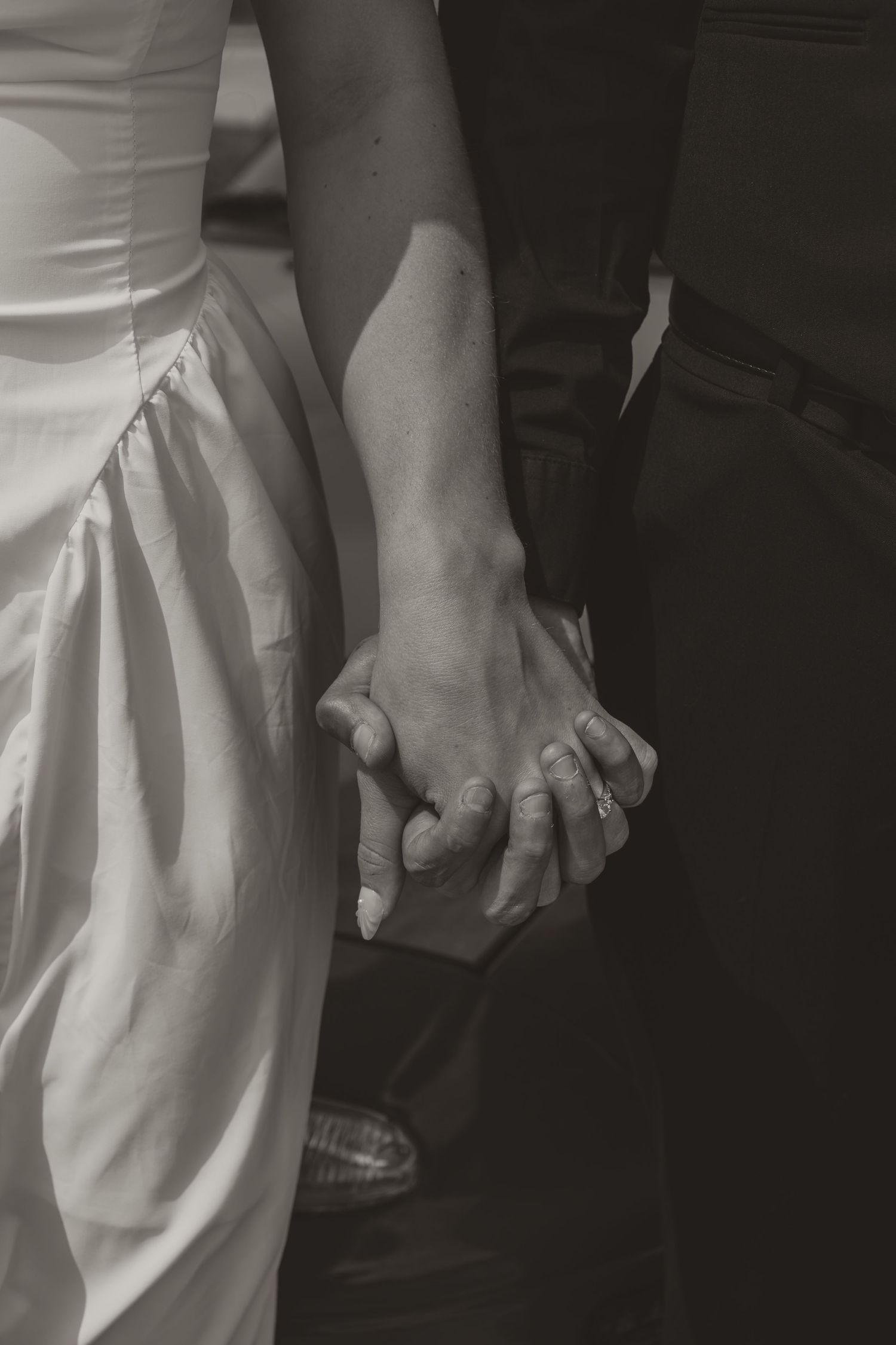 Close-up black and white photo of bride and groom holding hands, wedding rings visible.
