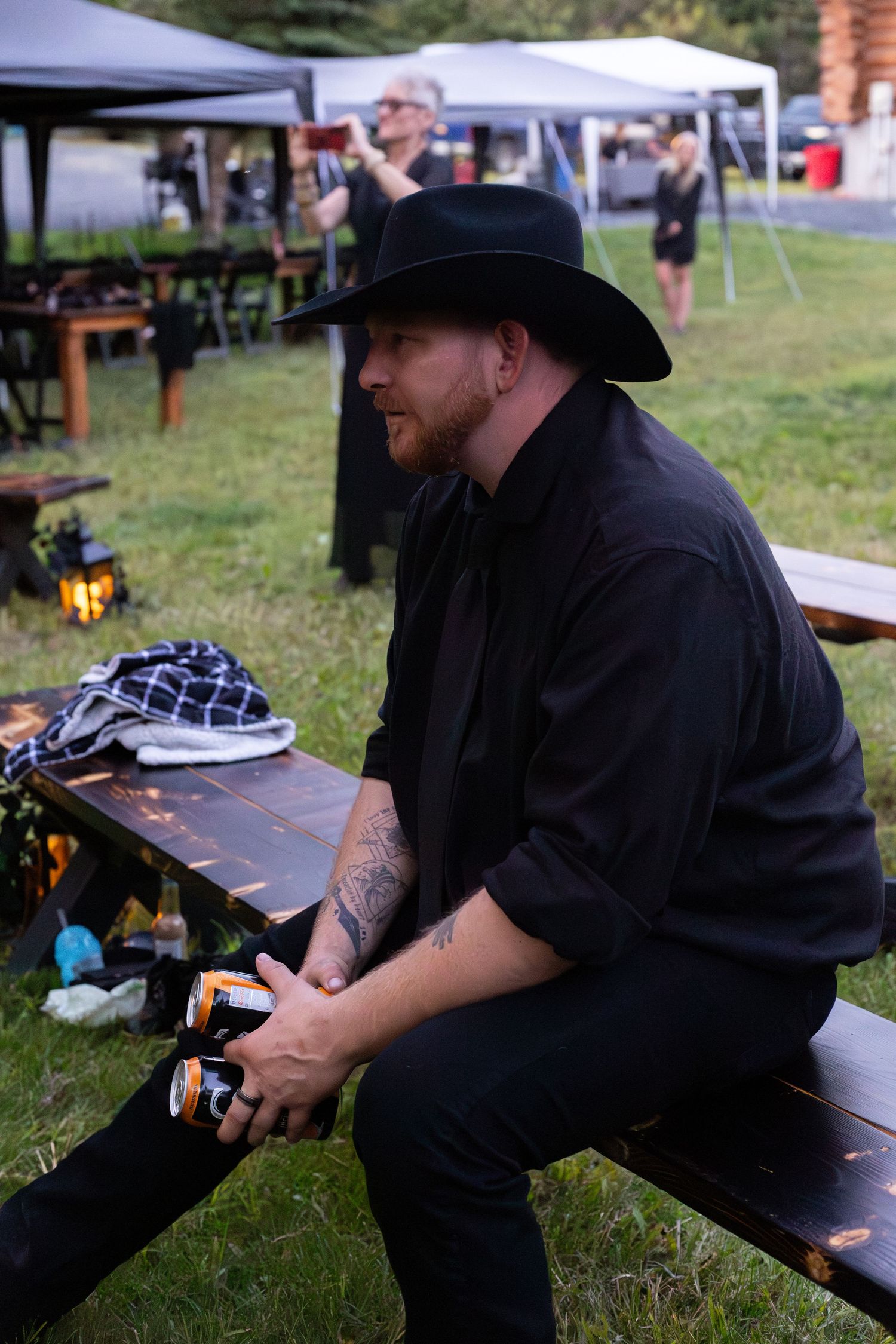 Man in black cowboy hat and shirt sitting on wooden bench at outdoor evening wedding reception.