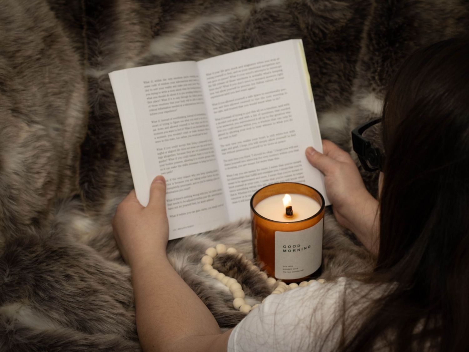 Person reading a book beside a lit soy candle on a cozy fur blanket.
