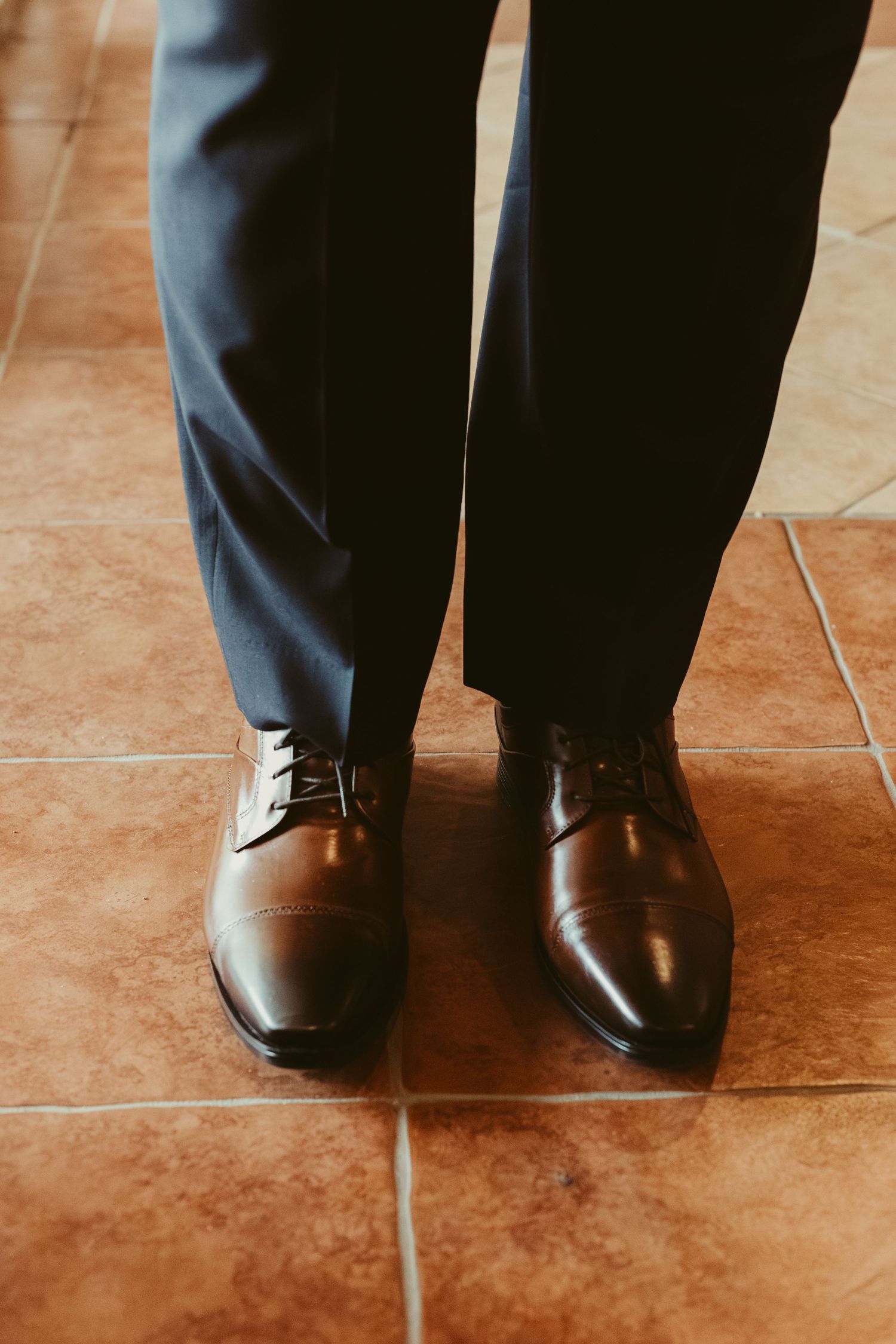 Groom wearing polished brown leather dress shoes and navy blue suit pants on tile floor.