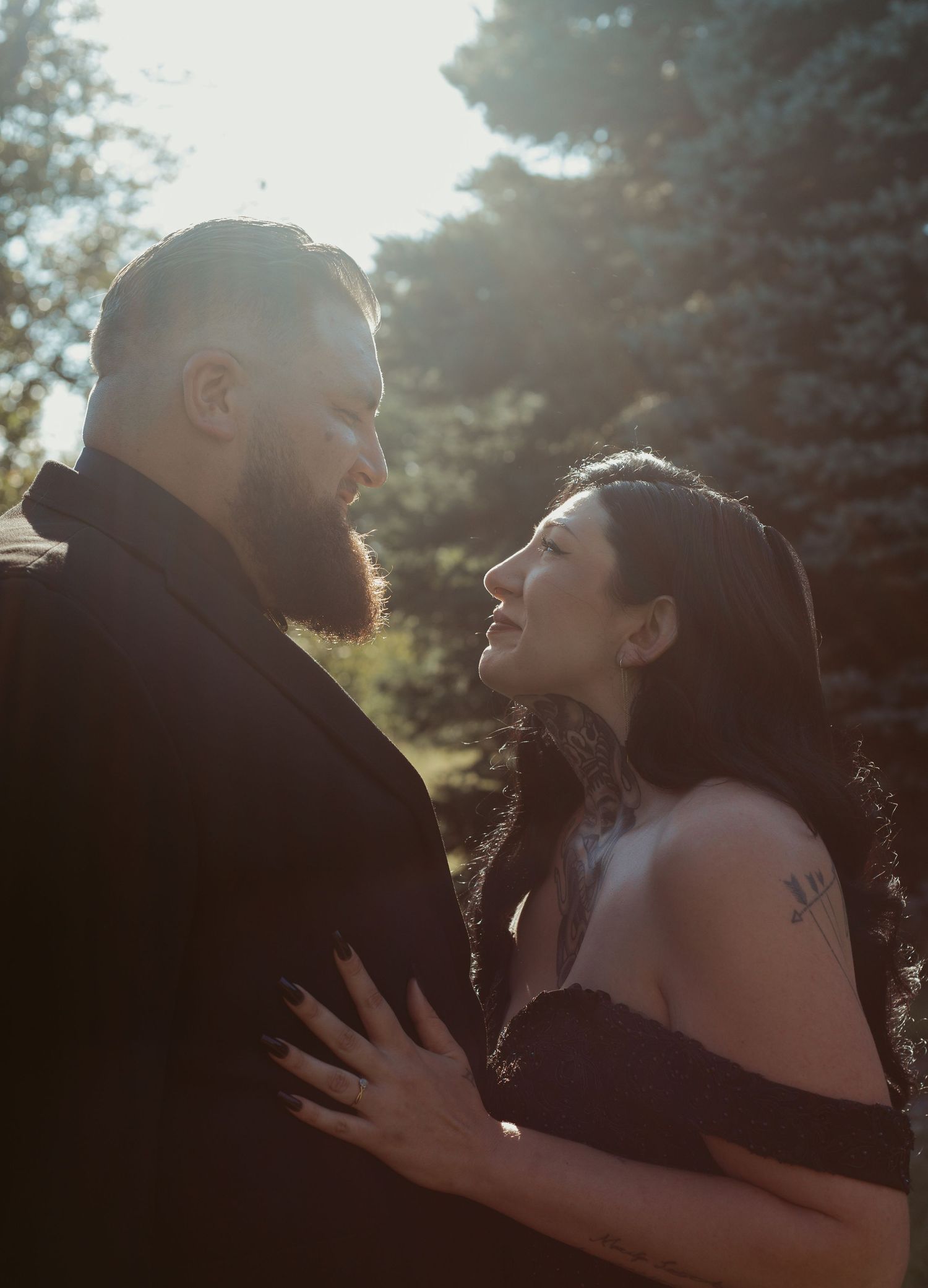 Bearded groom and tattooed bride gazing at each other in golden sunlight among pine trees.