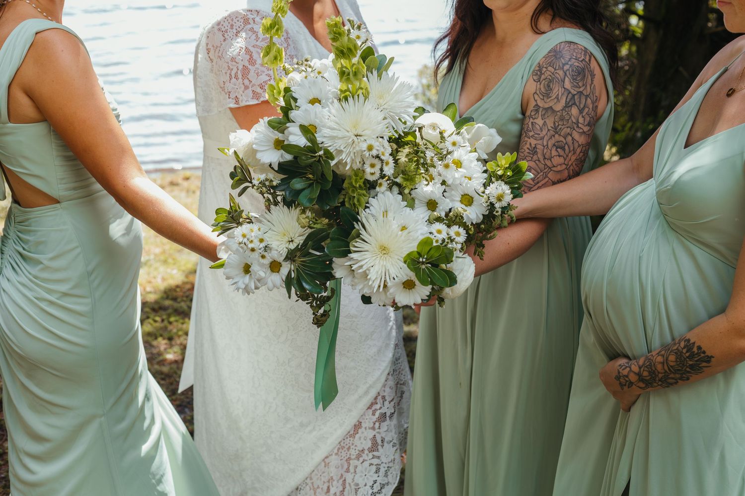 Close-up of white floral wedding bouquet held by bride and bridesmaids in sage green dresses outdoors.