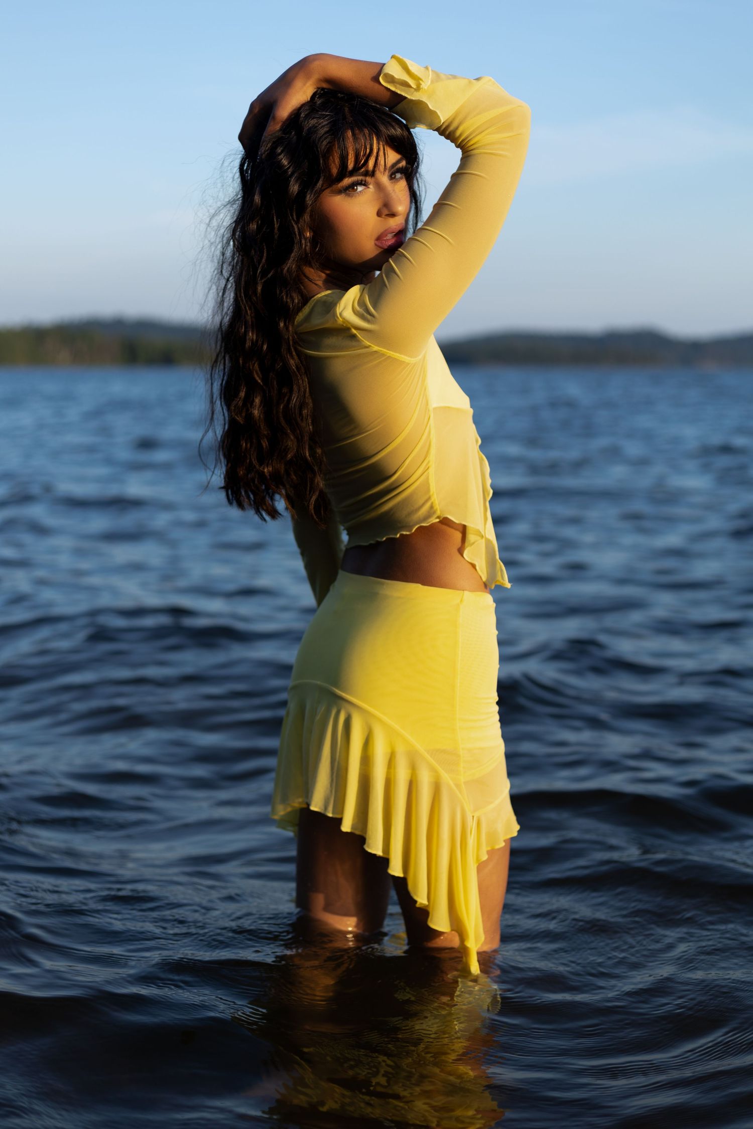 A young woman in a yellow crop top and ruffled skirt stands in a lake at golden hour.