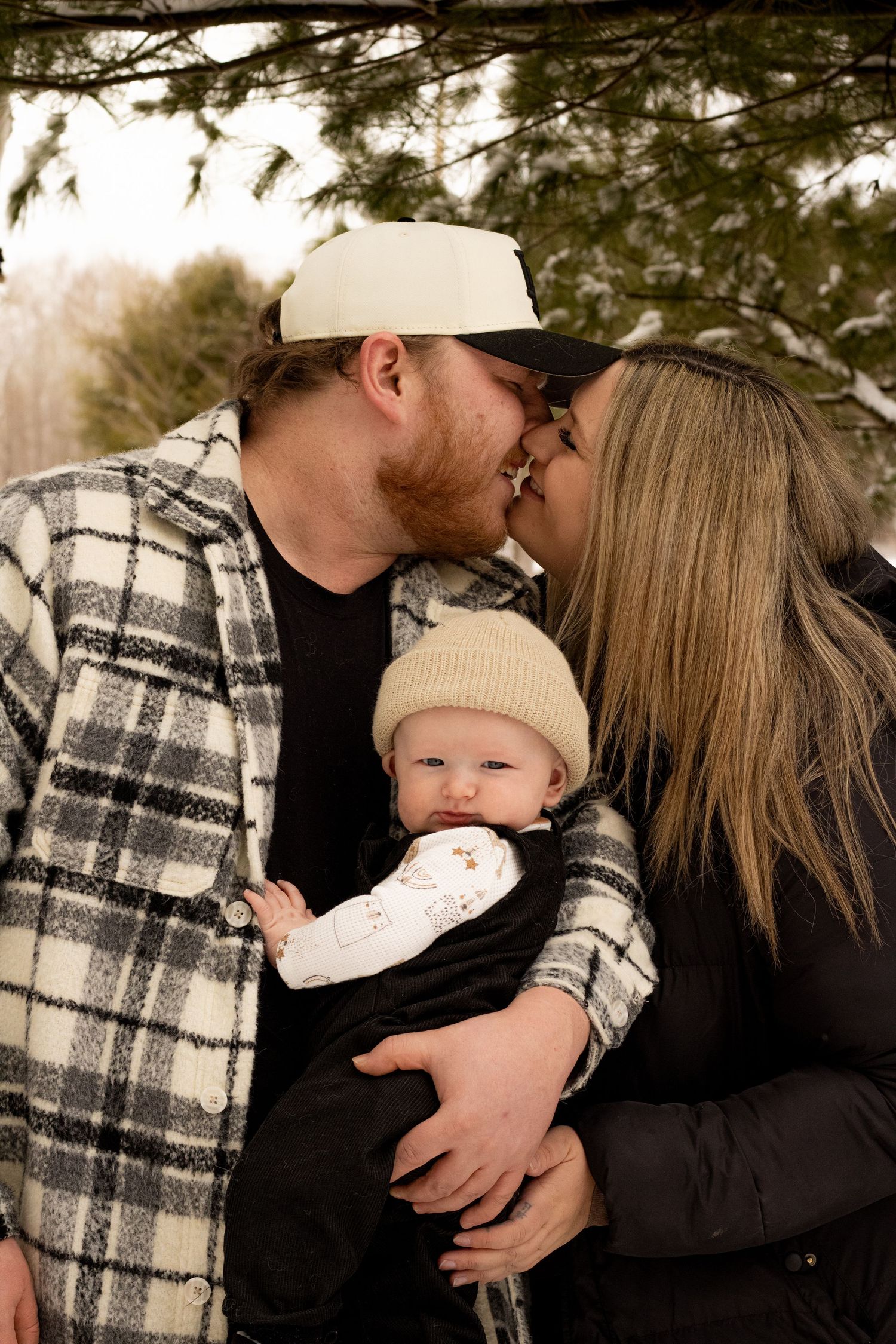 Couple kissing while holding their infant baby in a beanie hat outdoors in a snowy winter setting.