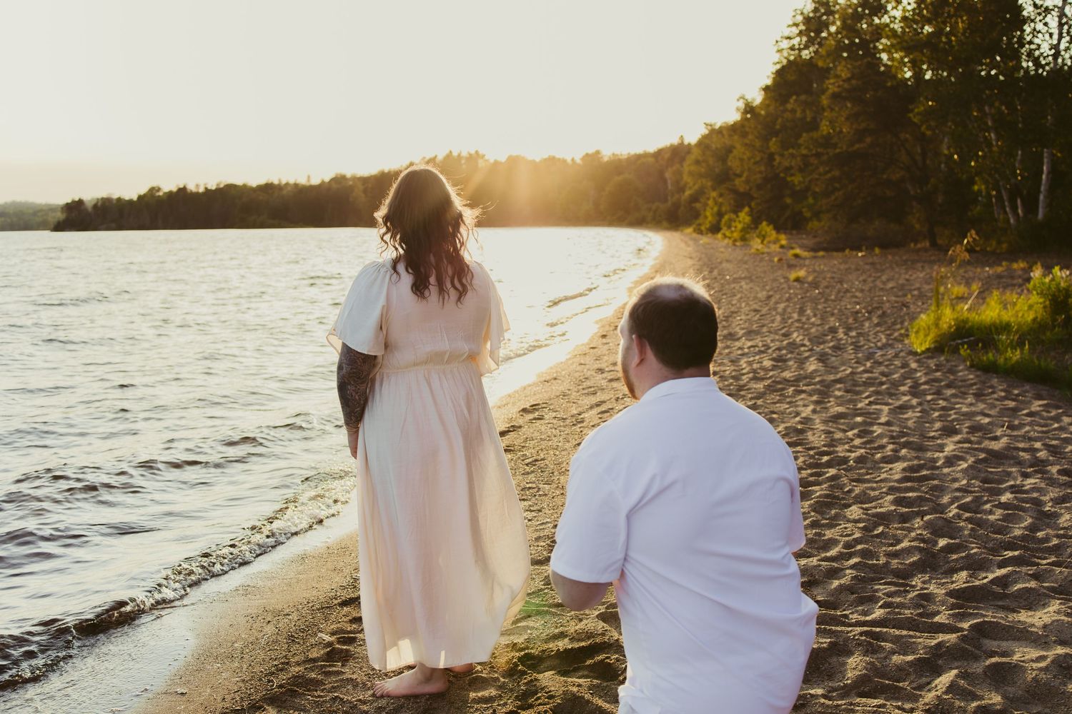 Couple walking hand in hand on a sandy beach at golden sunset with calm water and trees in background.