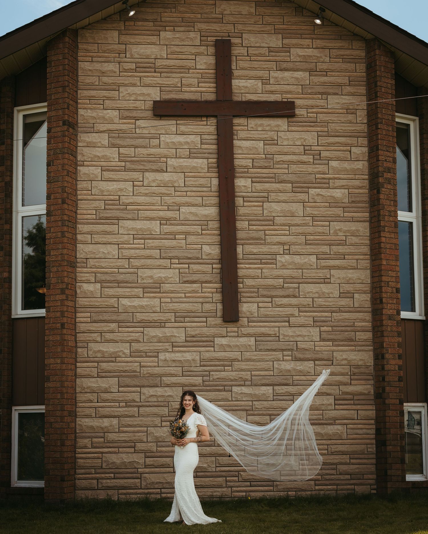 Bride in white wedding dress with flowing veil stands before a brick church wall with a large wooden cross.