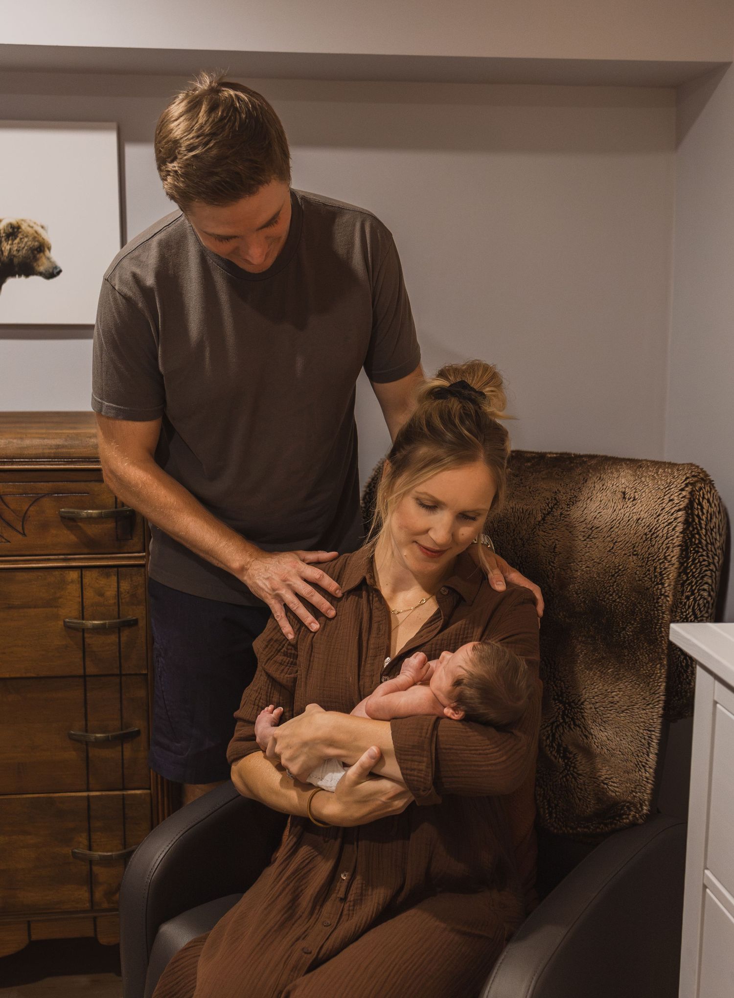New parents admire their newborn baby in a cozy nursery, mother seated in leather chair cradling infant.