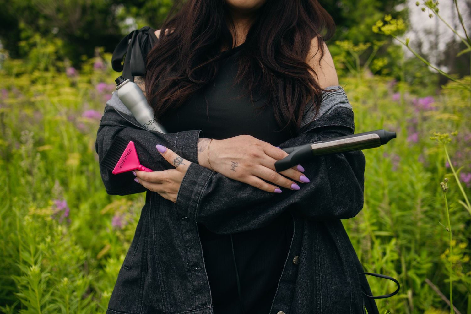Woman in black jacket holding pink and black Dyson hair tools outdoors among green and purple wildflowers.
