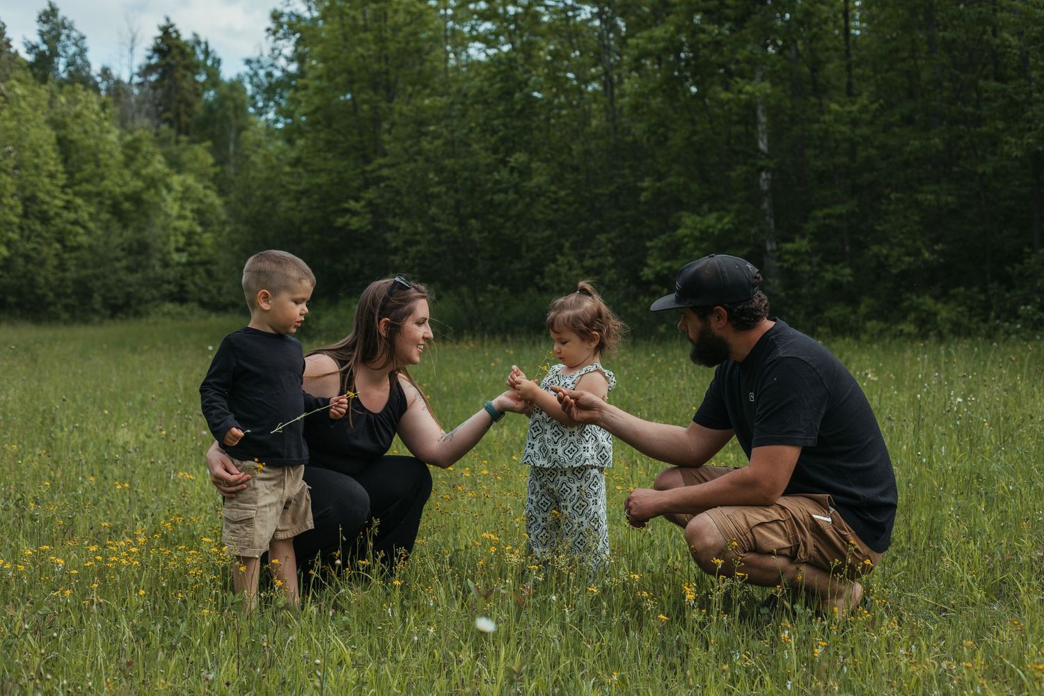 Family of four enjoying a sunny day in a lush green meadow surrounded by trees, sharing a tender moment together.