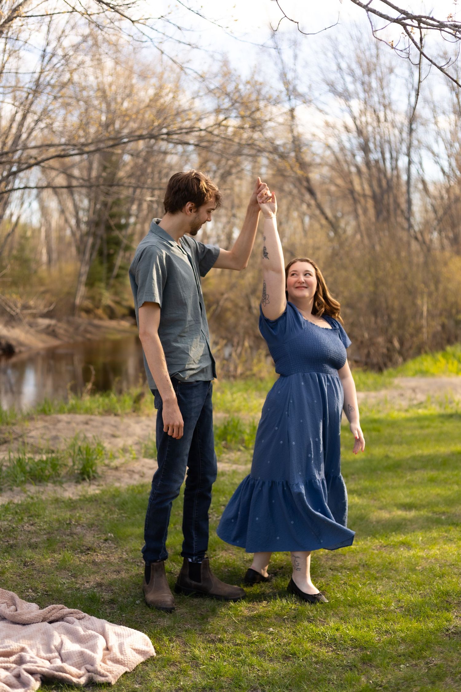 Couple dancing outdoors in spring, woman in blue dress spins as man holds her hand near a peaceful creek.