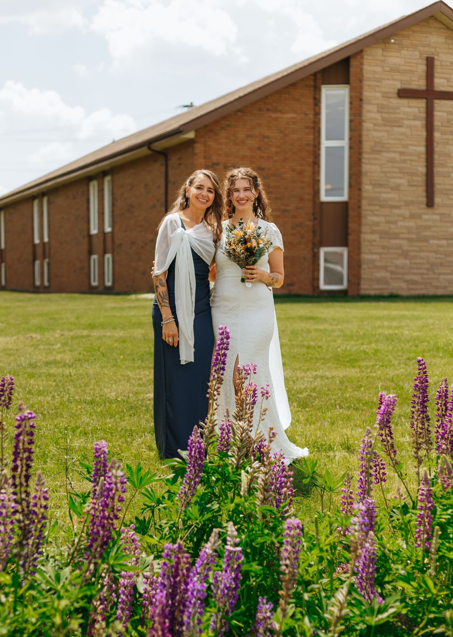 Bride in white gown and bridesmaid in navy dress pose outside brick church with purple lupine flowers.