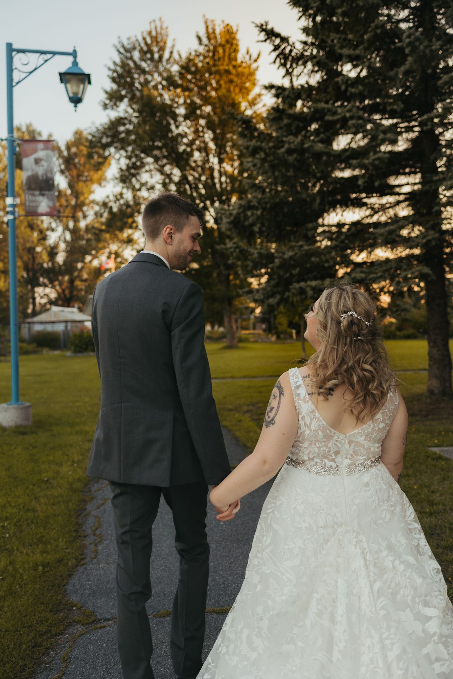 Bride and groom walking hand in hand through a park at sunset, bride in lace gown, groom in dark suit.