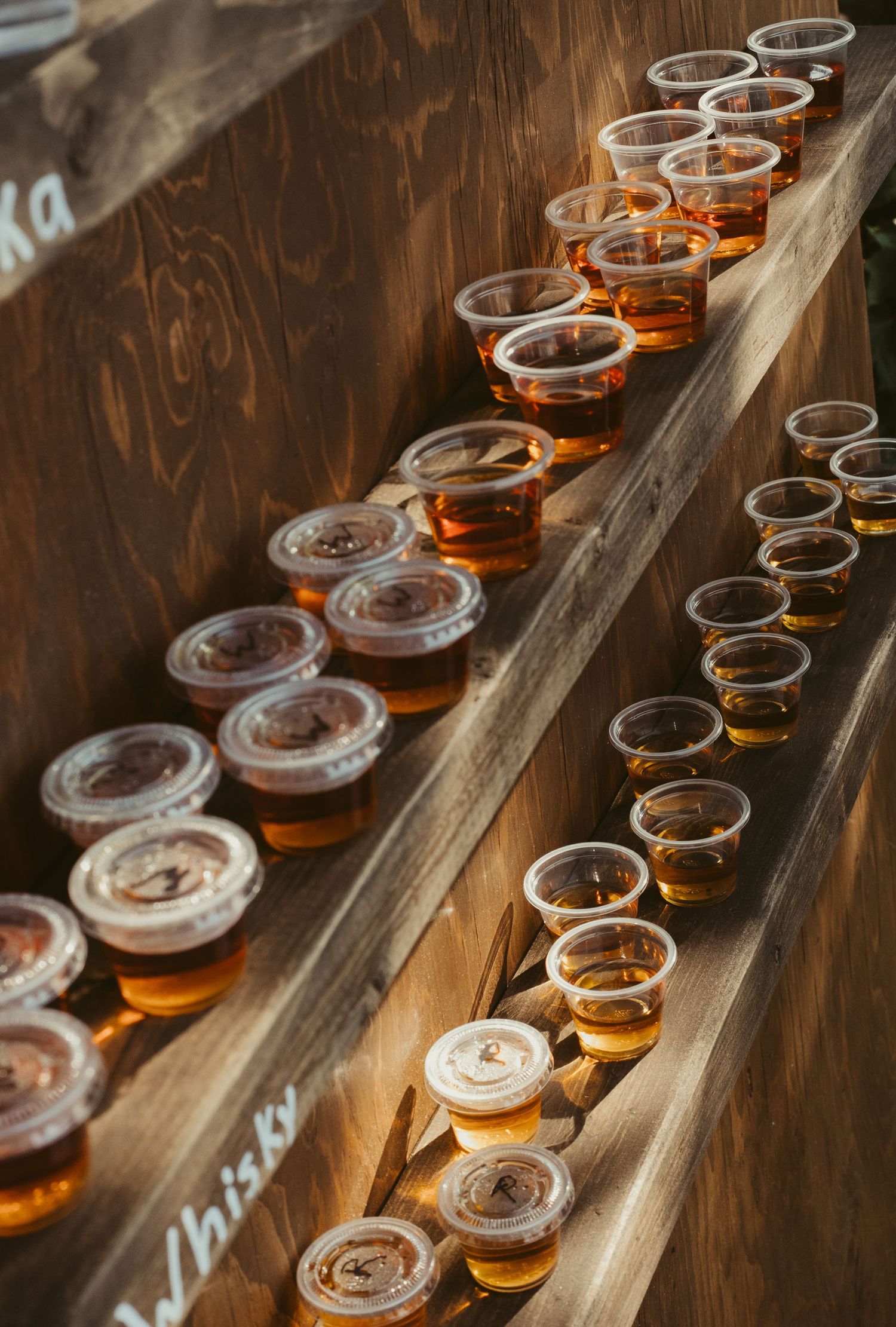 Rows of whiskey shot cups lined up on rustic wooden shelves at an outdoor wedding reception.