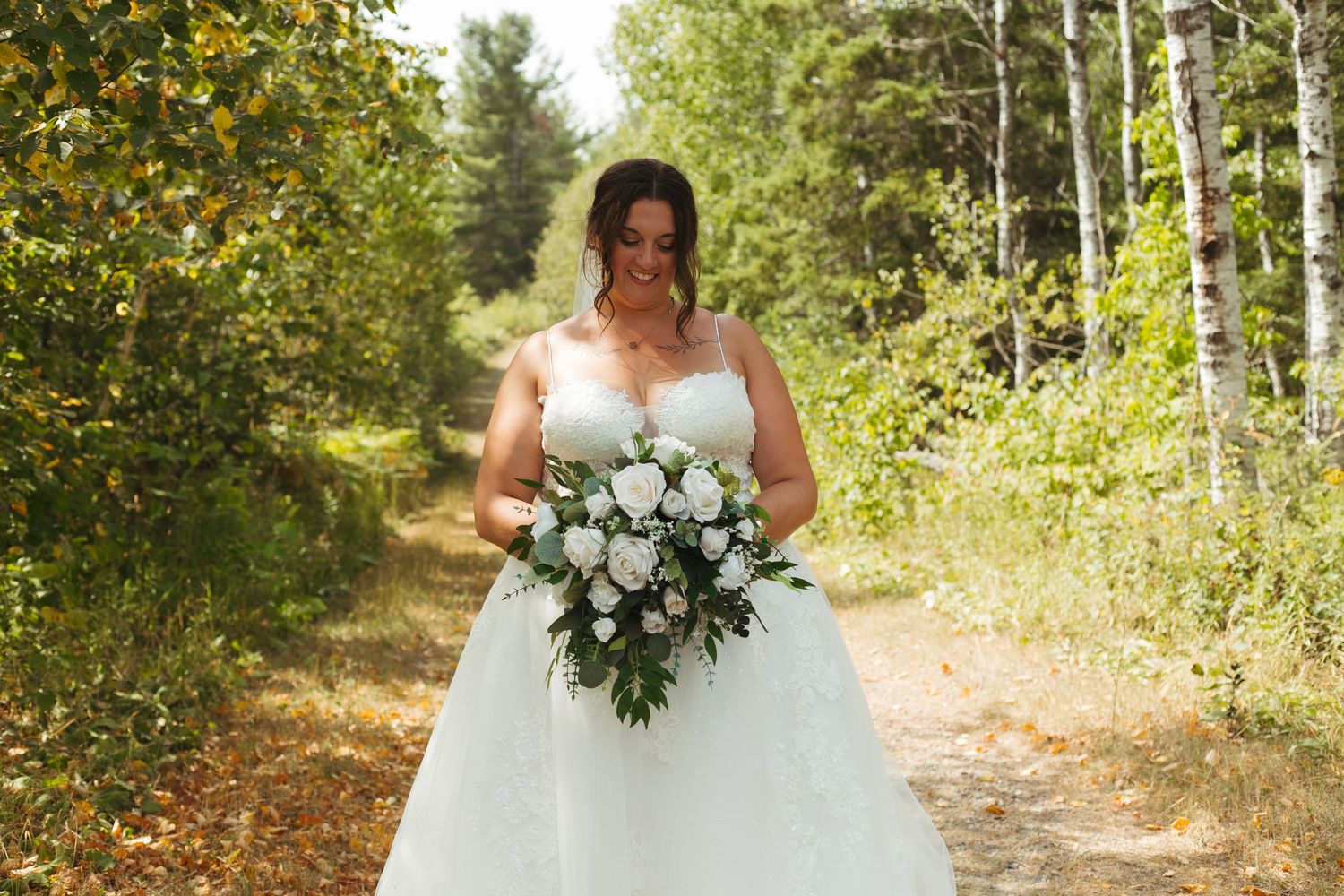 Bride in strapless ball gown holding white bouquet walking down forest path.