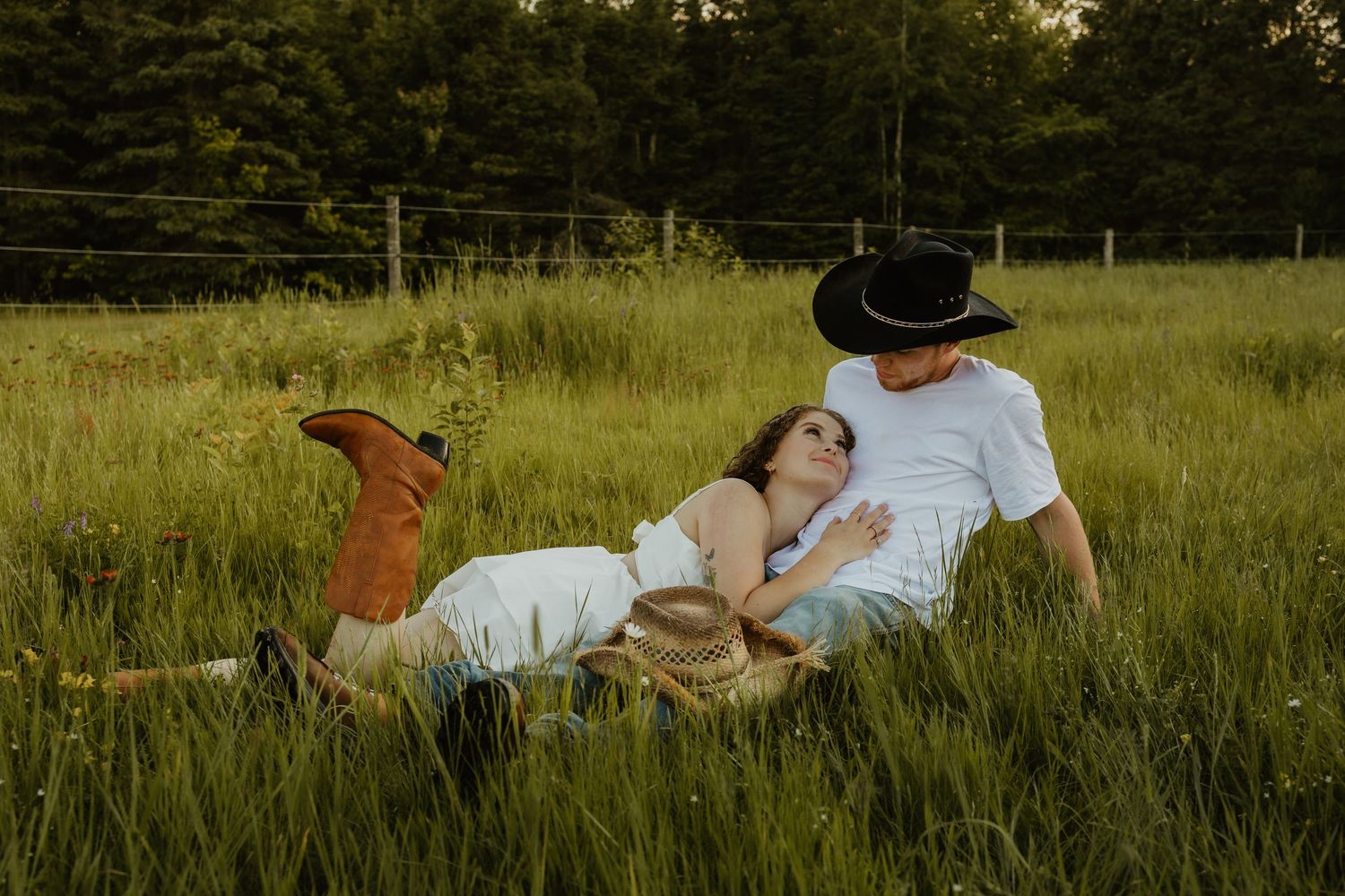 Couple relaxing in lush green field, woman in cowboy boots lying on mans lap, both wearing hats, romantic outdoor photo.
