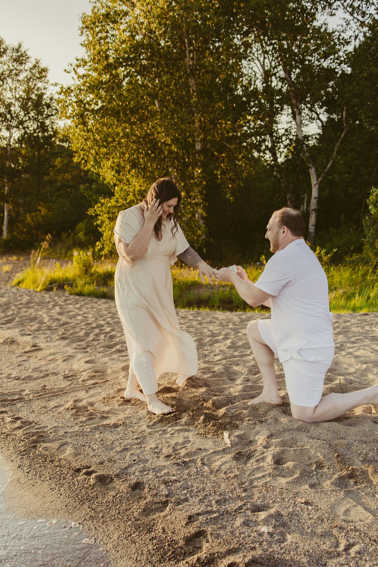 Man proposes on sandy beach at sunset, kneeling before woman in cream dress, with lush green trees in background.