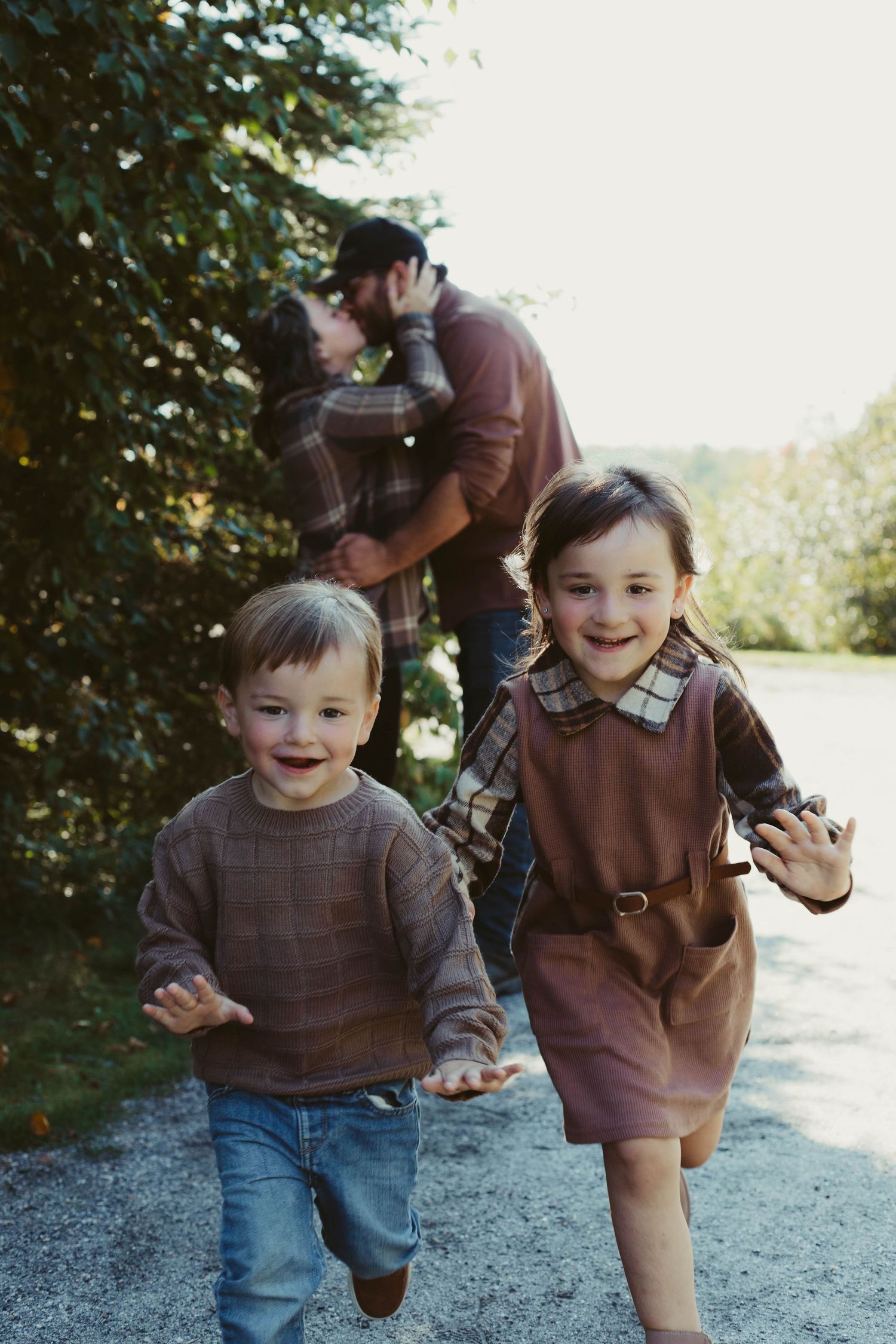 Two young children run toward camera while parents embrace near evergreen trees.