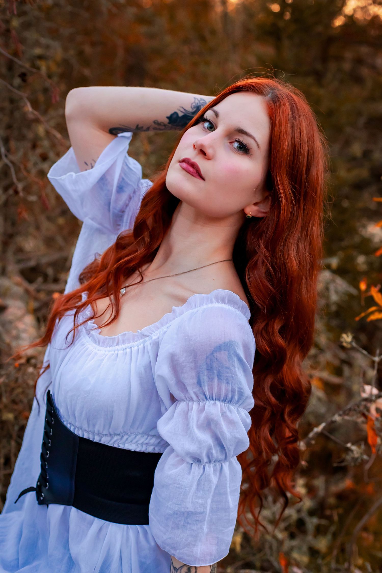 Striking woman with long red hair in white off-shoulder dress and black corset in autumn forest.