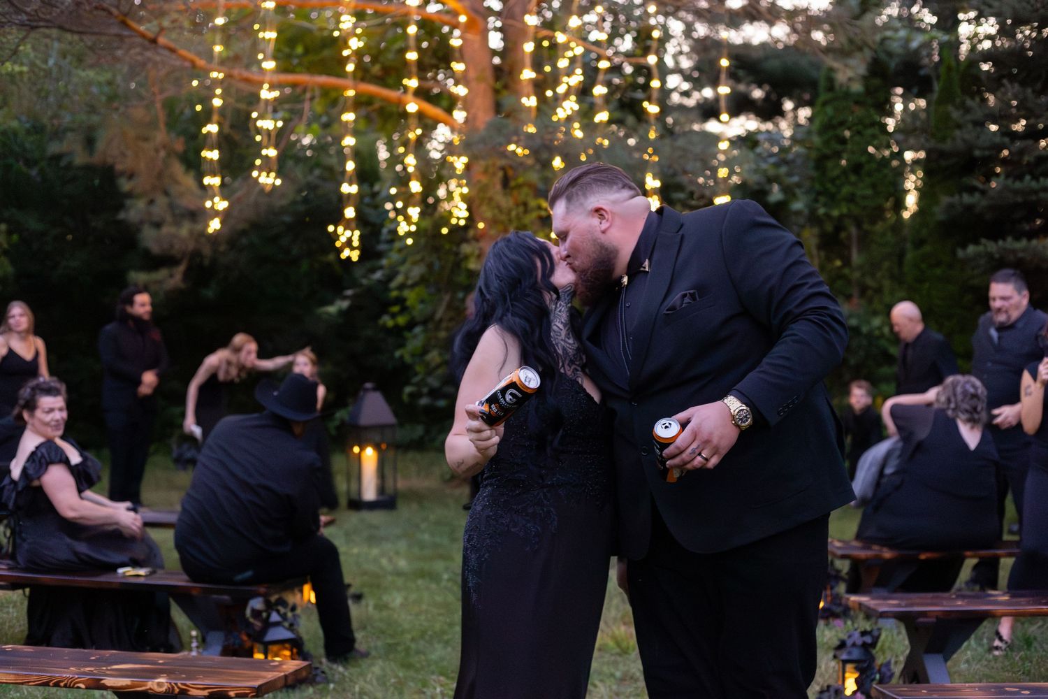 Newlywed couple sharing first kiss at outdoor gothic wedding with guests and string lights in background.