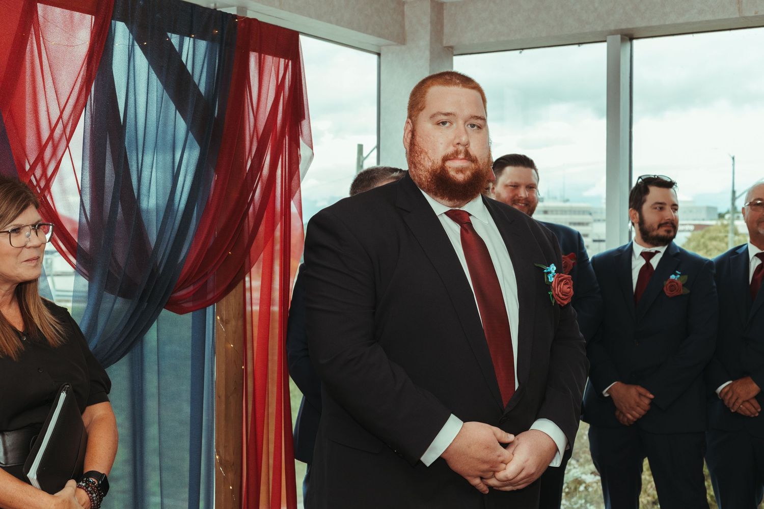 Groom in black suit with burgundy tie standing at altar with groomsmen in navy suits behind him.