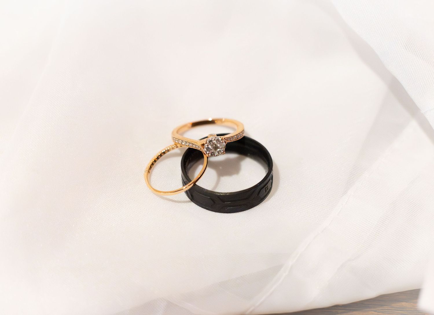 Gold diamond engagement ring and black wedding band resting together on white fabric.