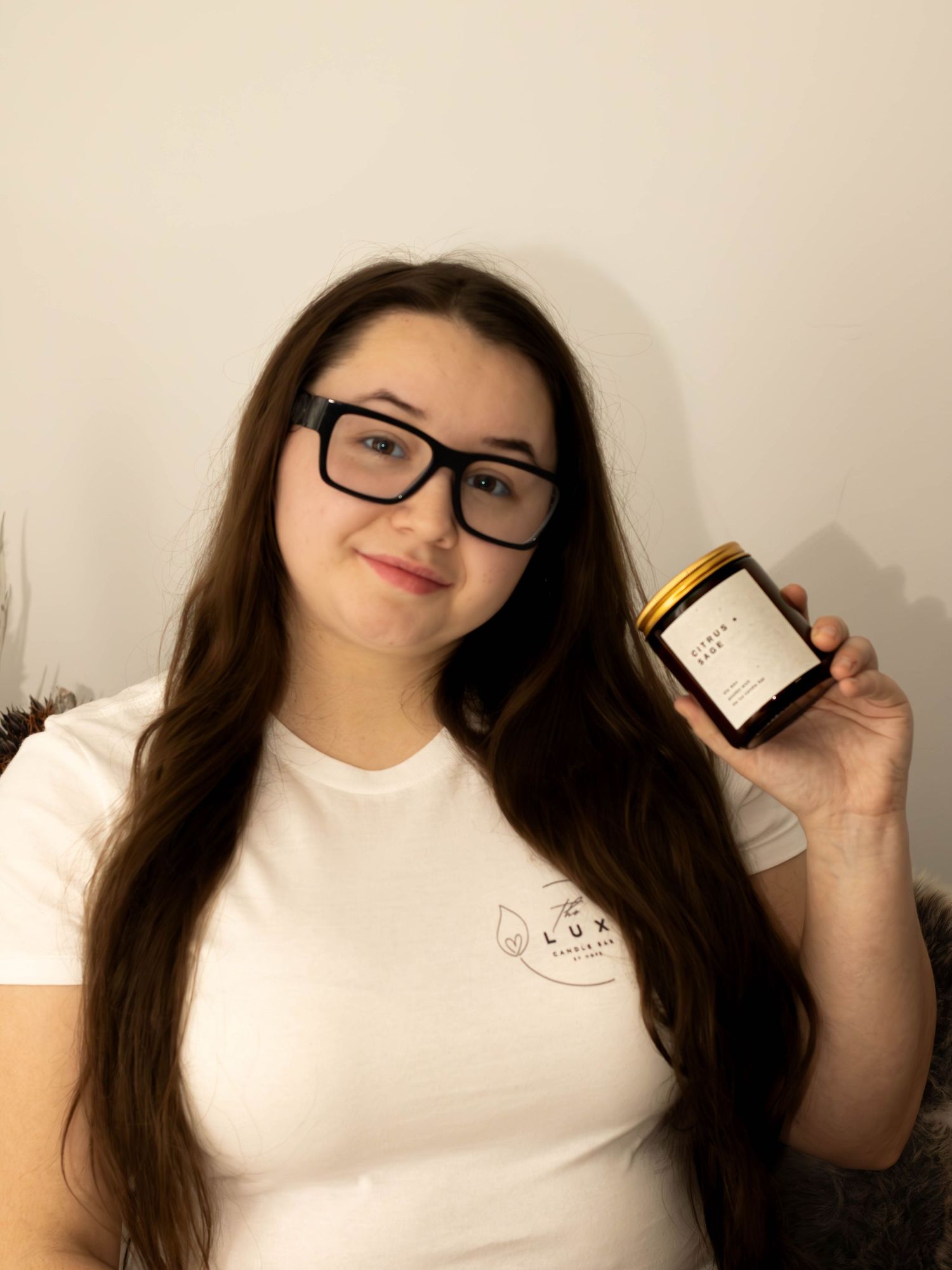 Young woman with glasses holding a gold-lidded candle jar wearing a white Lux branded t-shirt.