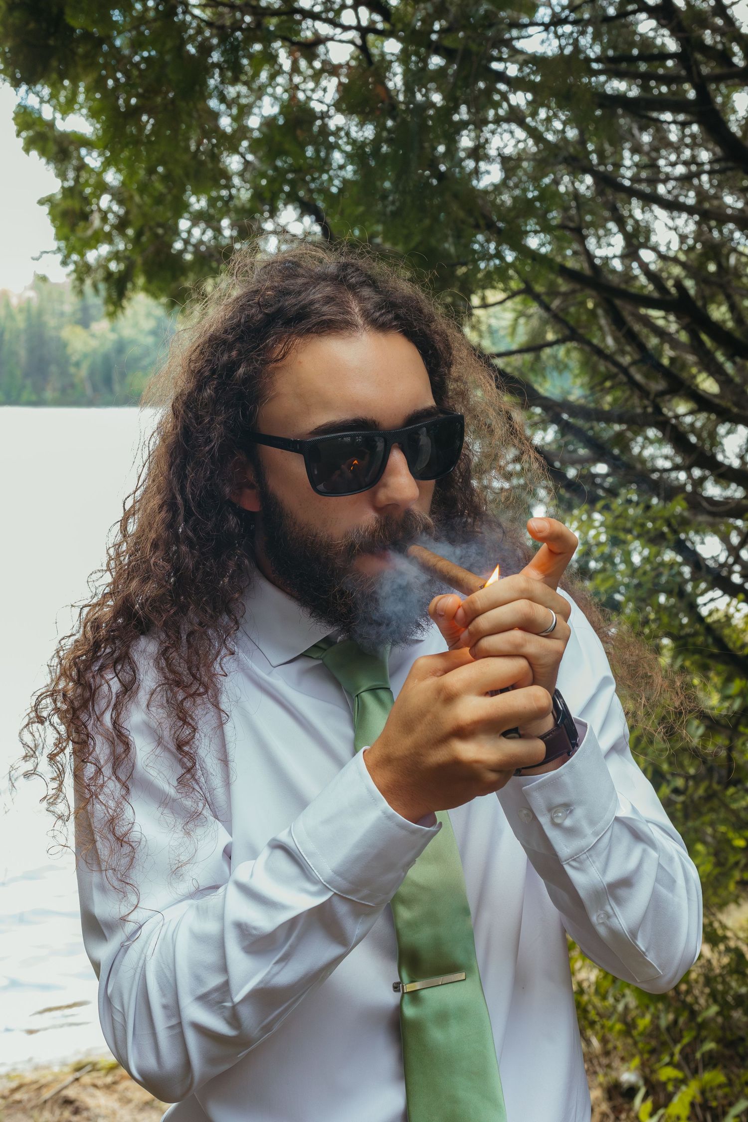 Long-haired groom with sunglasses lighting a cigar outdoors near a lake on his wedding day.
