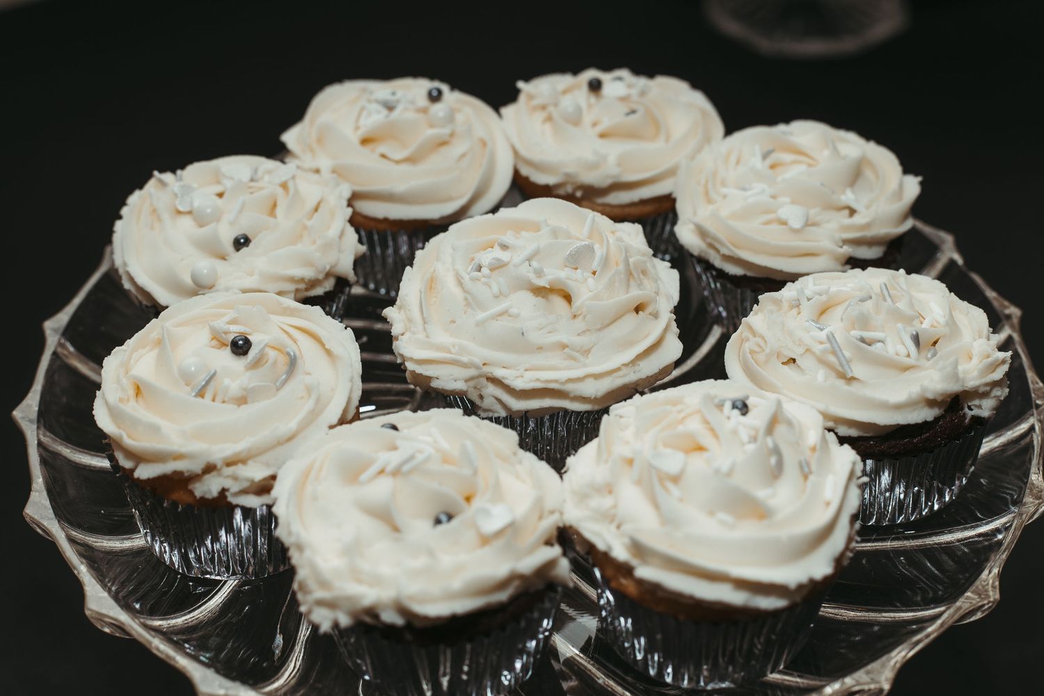 Nine chocolate cupcakes with swirled white frosting and silver pearl decorations on glass cake stand.