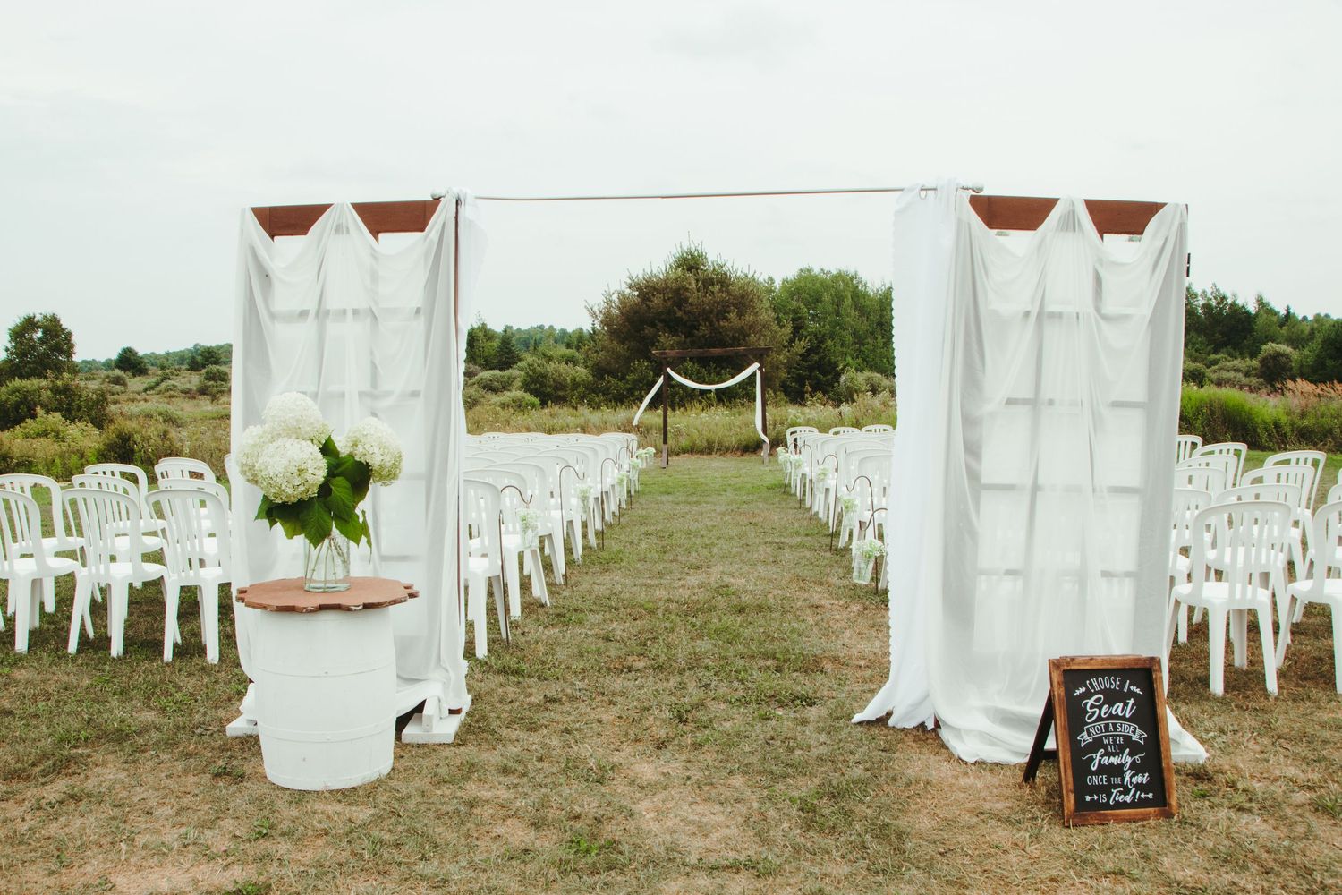 An elegant outdoor wedding aisle with white draping arch, chairs, and a chalkboard sign.