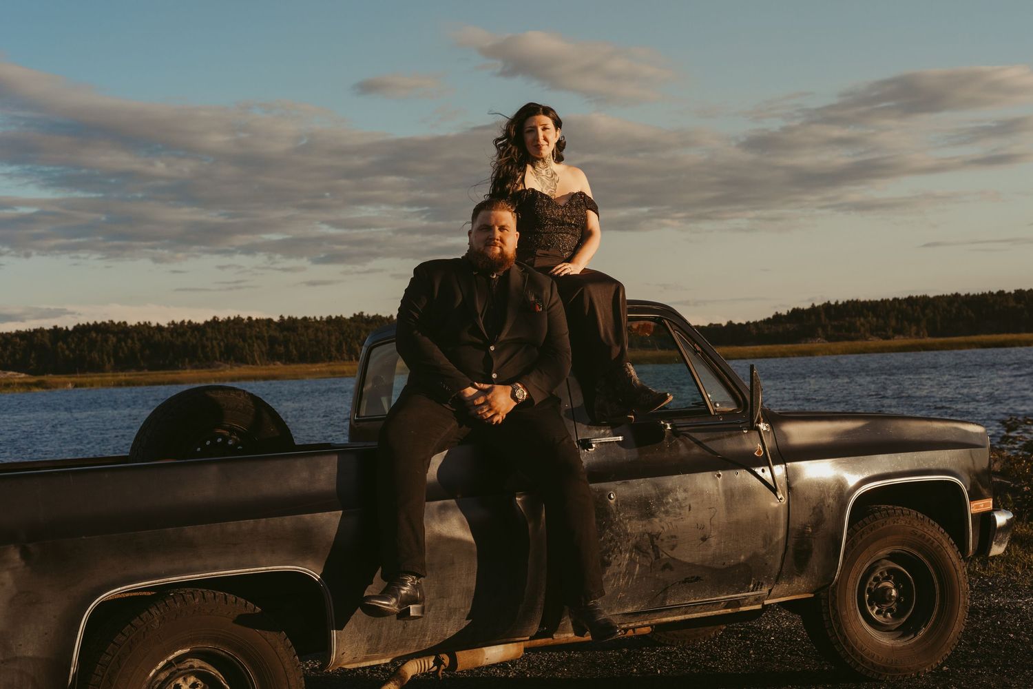 Couple posing on hood of a rustic vintage pickup truck at golden hour beside a scenic waterway.
