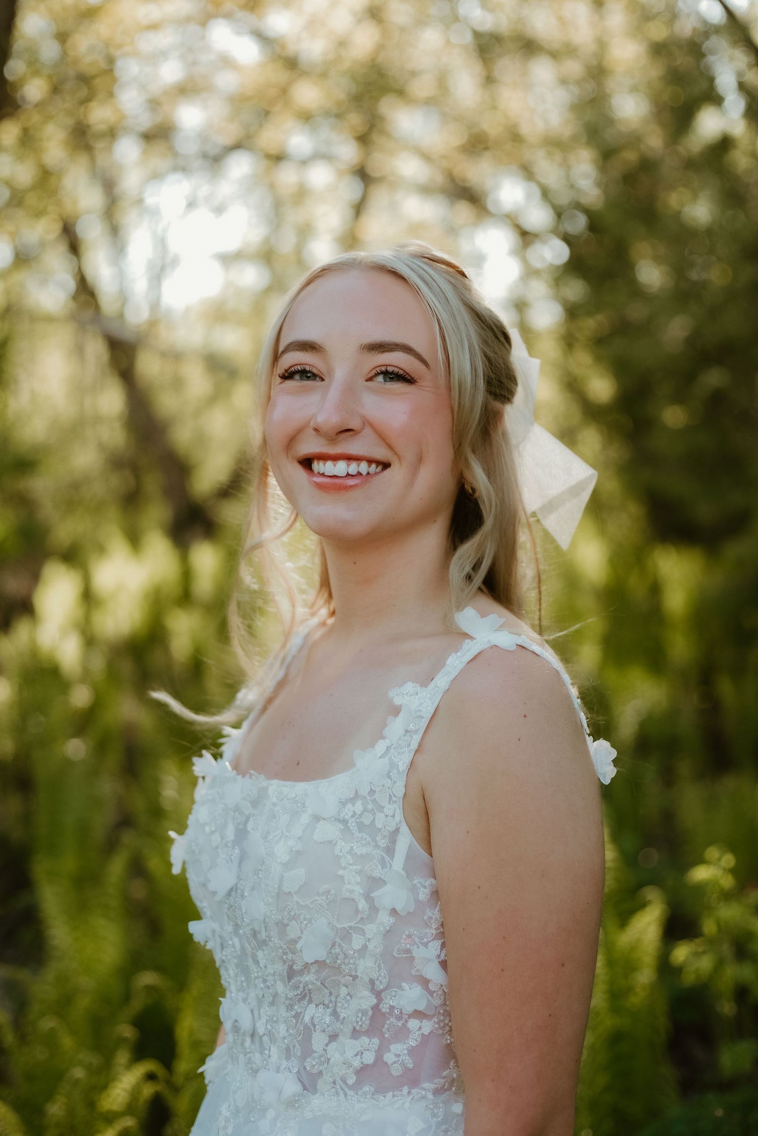Smiling blonde bride in white lace floral gown with bow hair accessory, posing outdoors in lush green nature setting.