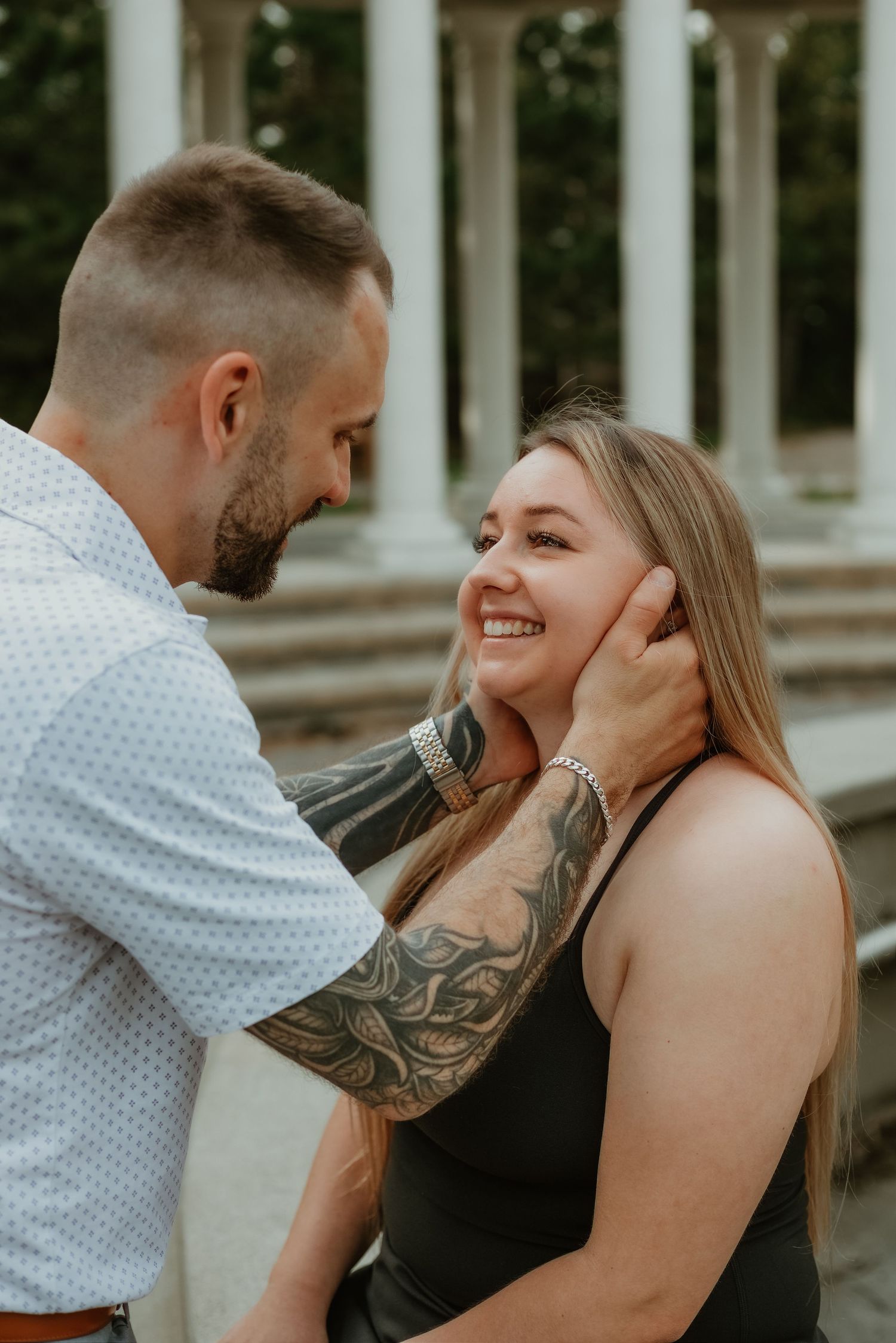 A tattooed man tenderly cups a smiling woman's face near elegant stone columns outdoors.