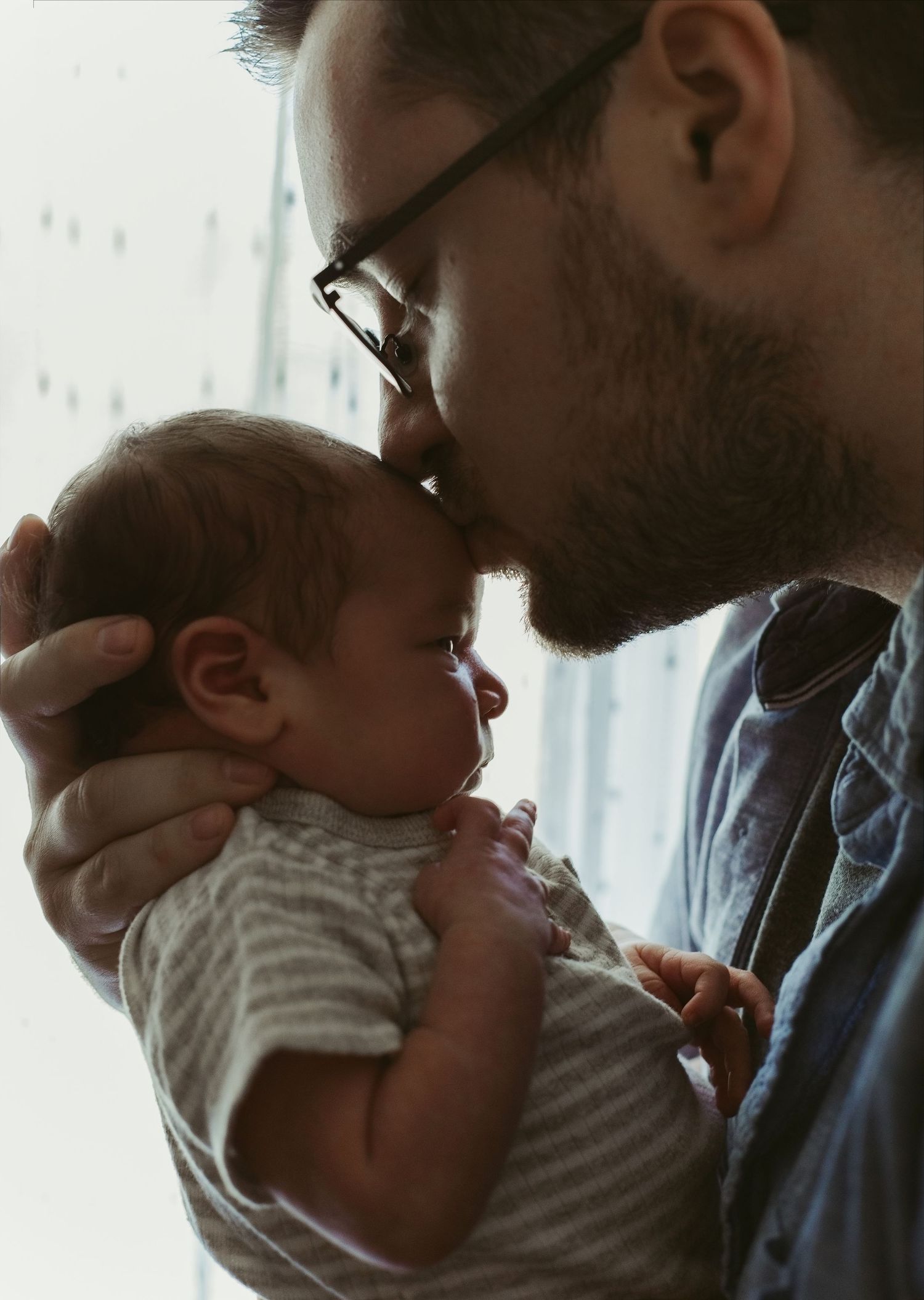 A bearded father wearing glasses tenderly kisses his newborn baby on the forehead while cradling the infant.