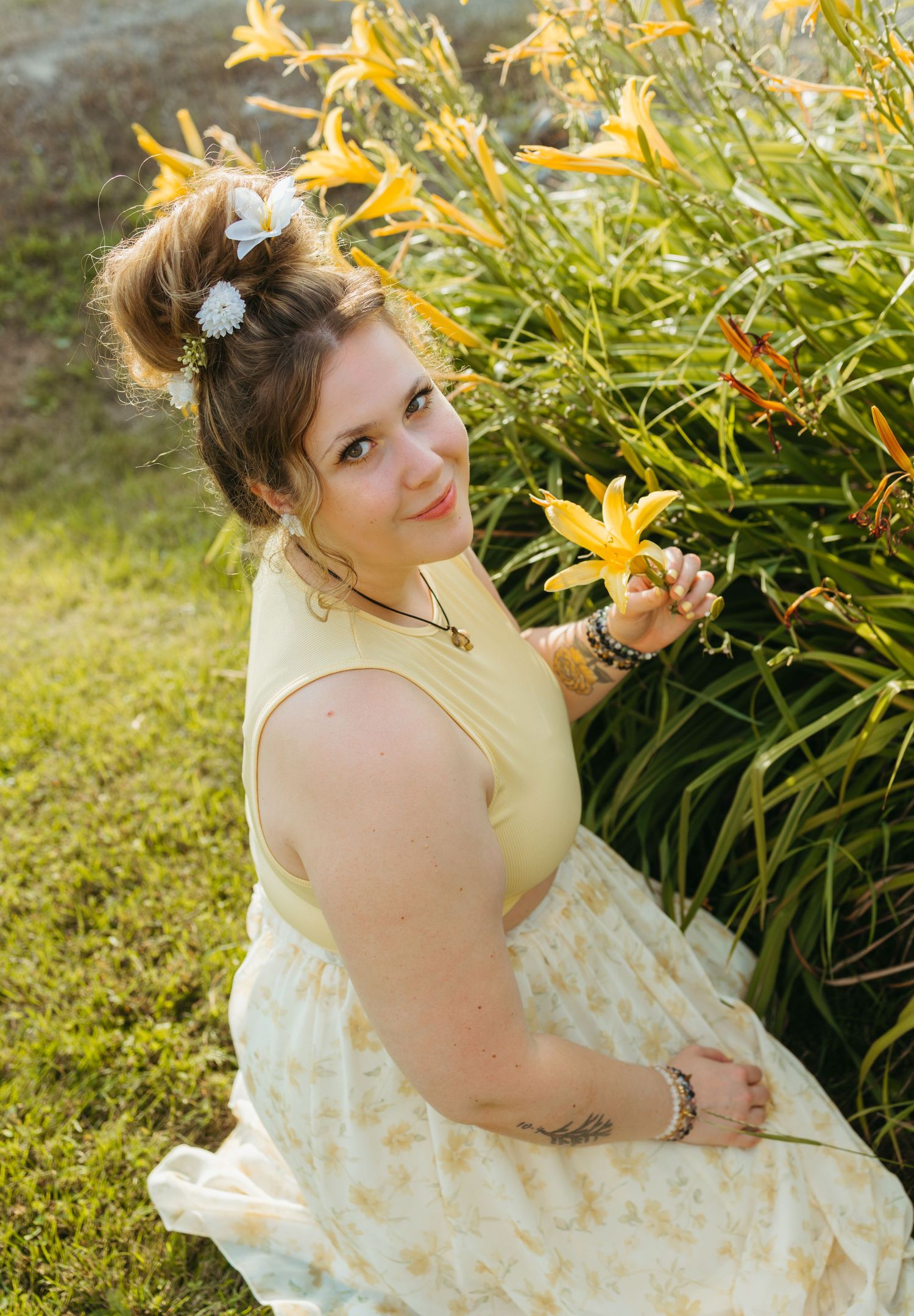 Young woman in yellow floral dress holding orange lily beside garden flowers with hair accessories outdoors.
