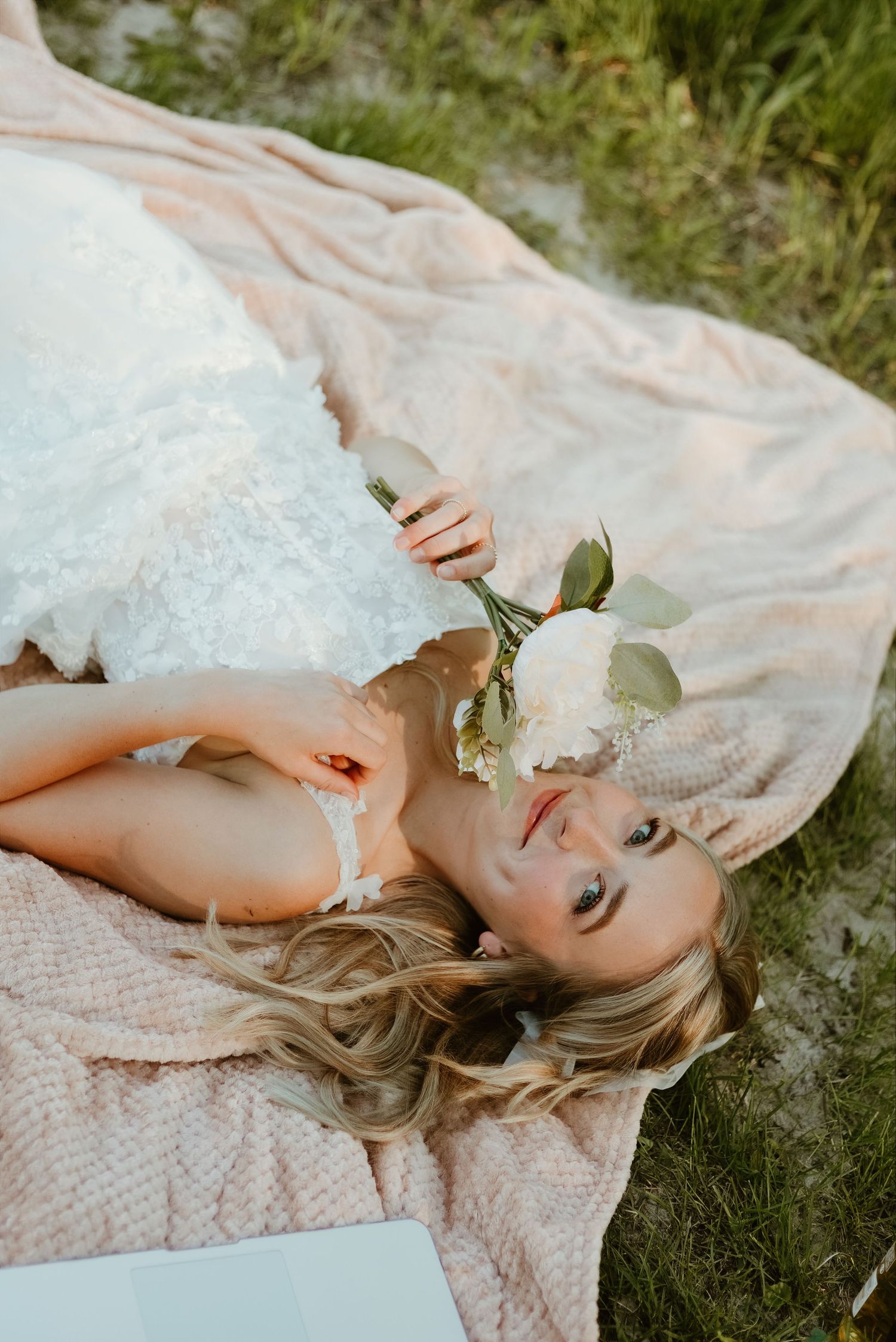 Bride in white lace gown lying on pink blanket holding white peony flowers outdoors.