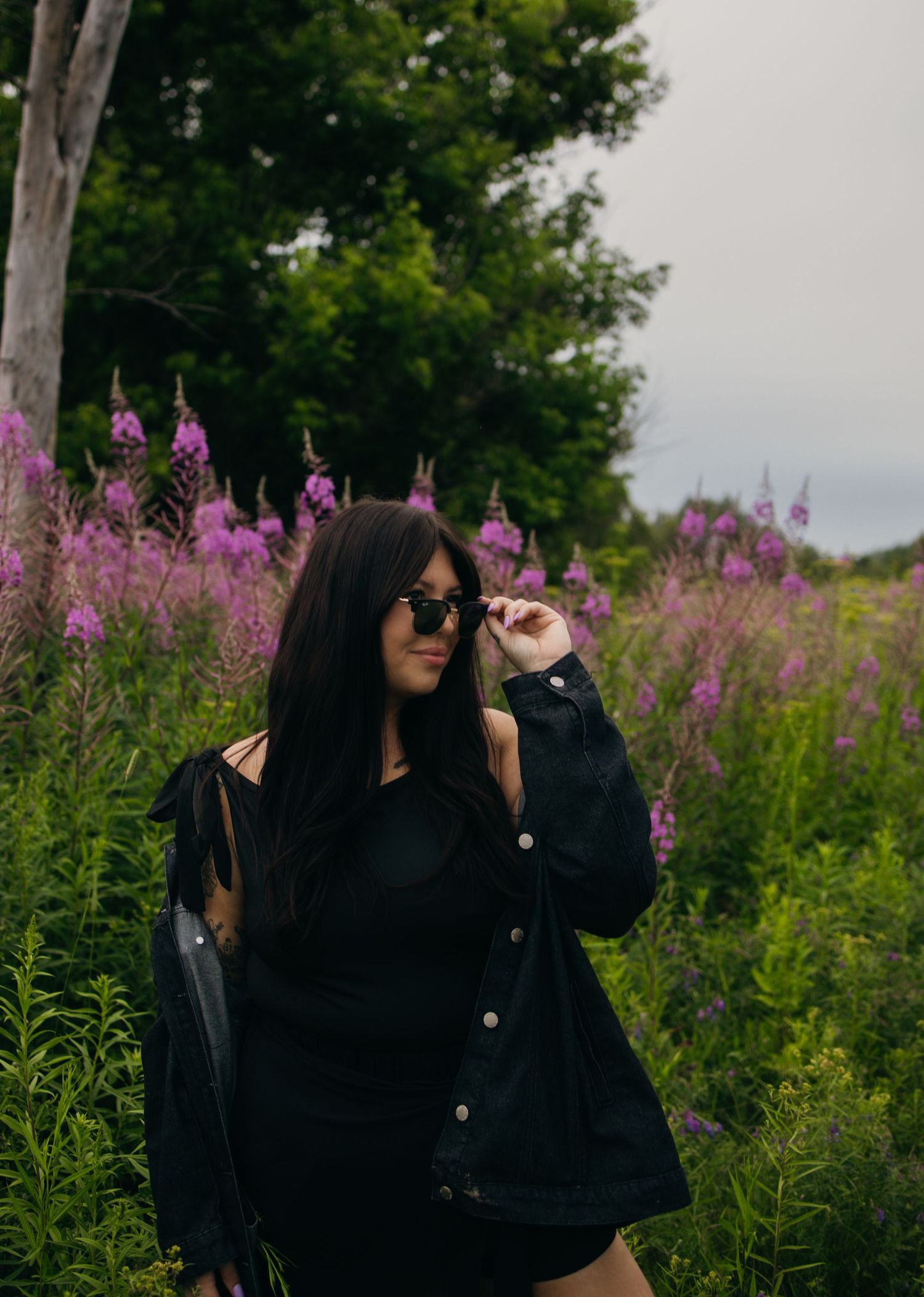 Woman in black outfit and sunglasses poses among vibrant purple wildflowers in a lush green field.
