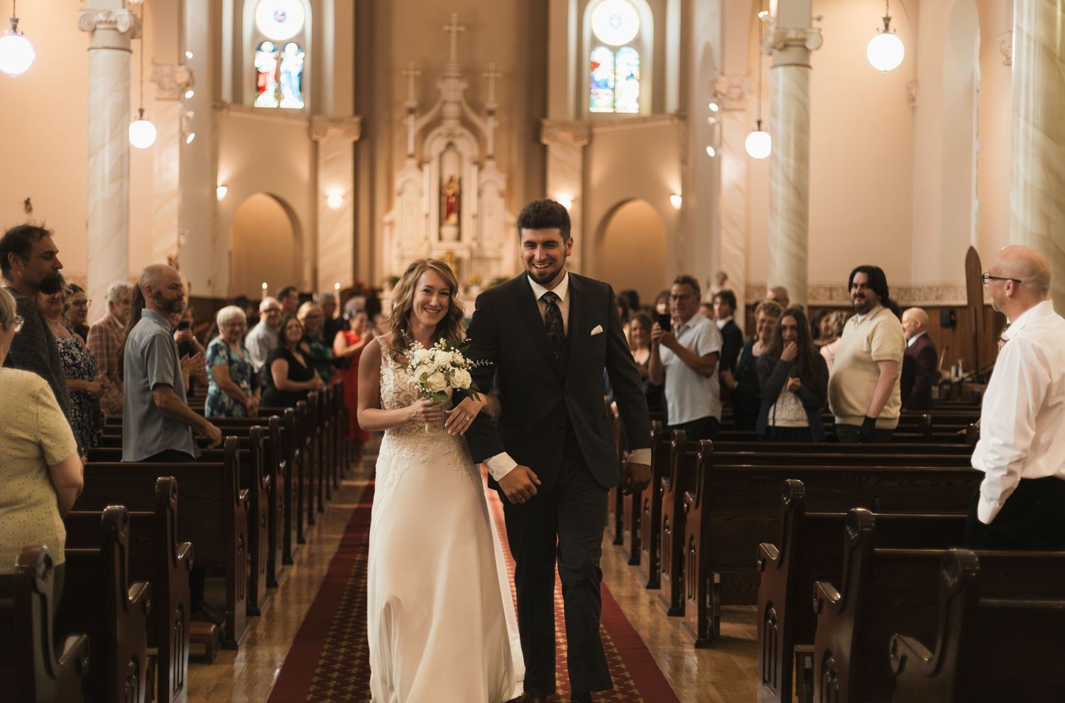 Newlywed couple walks down church aisle after wedding ceremony, surrounded by applauding guests in pews.