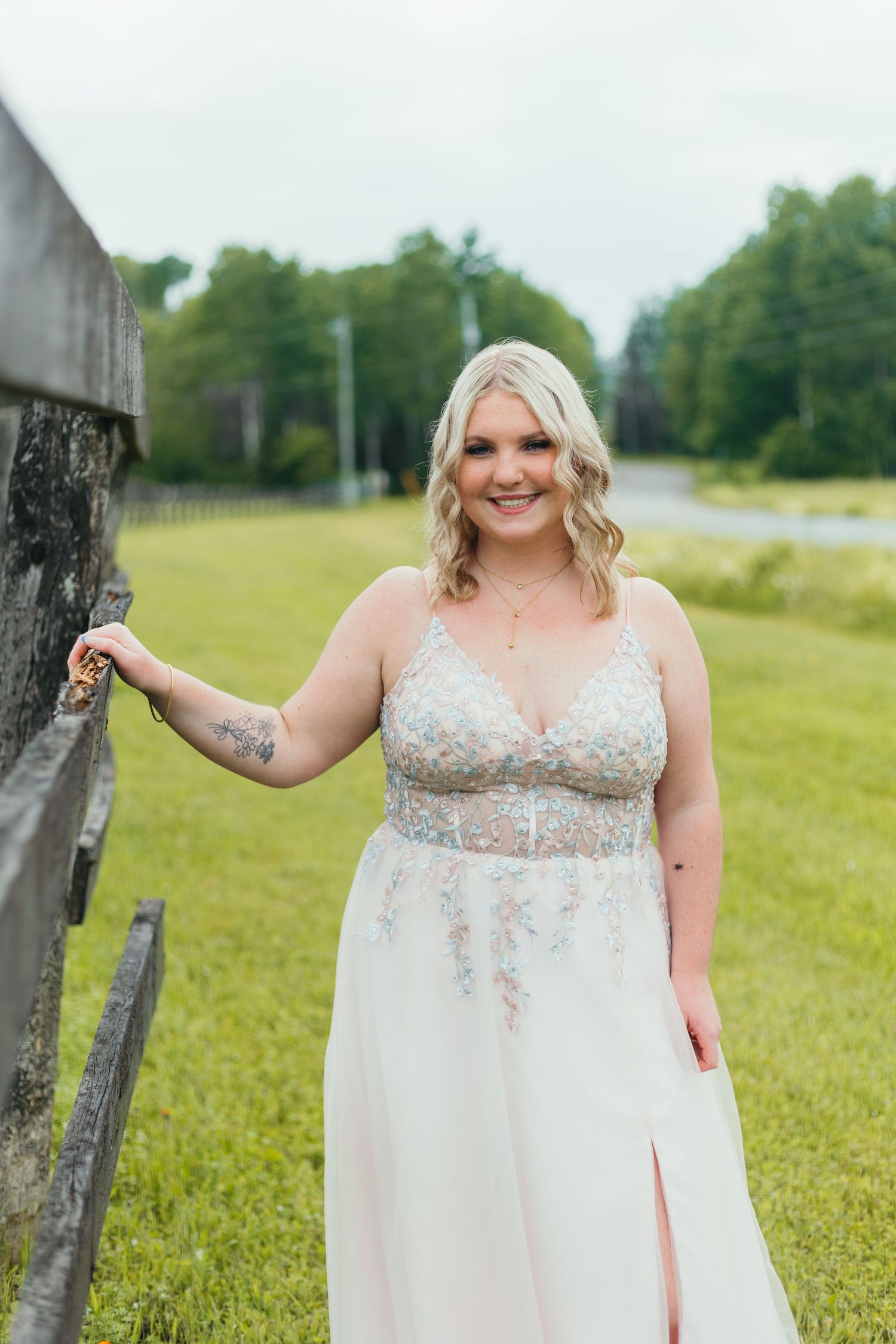 A blonde woman in a floral embroidered blush prom gown poses by a wooden fence in a green field.
