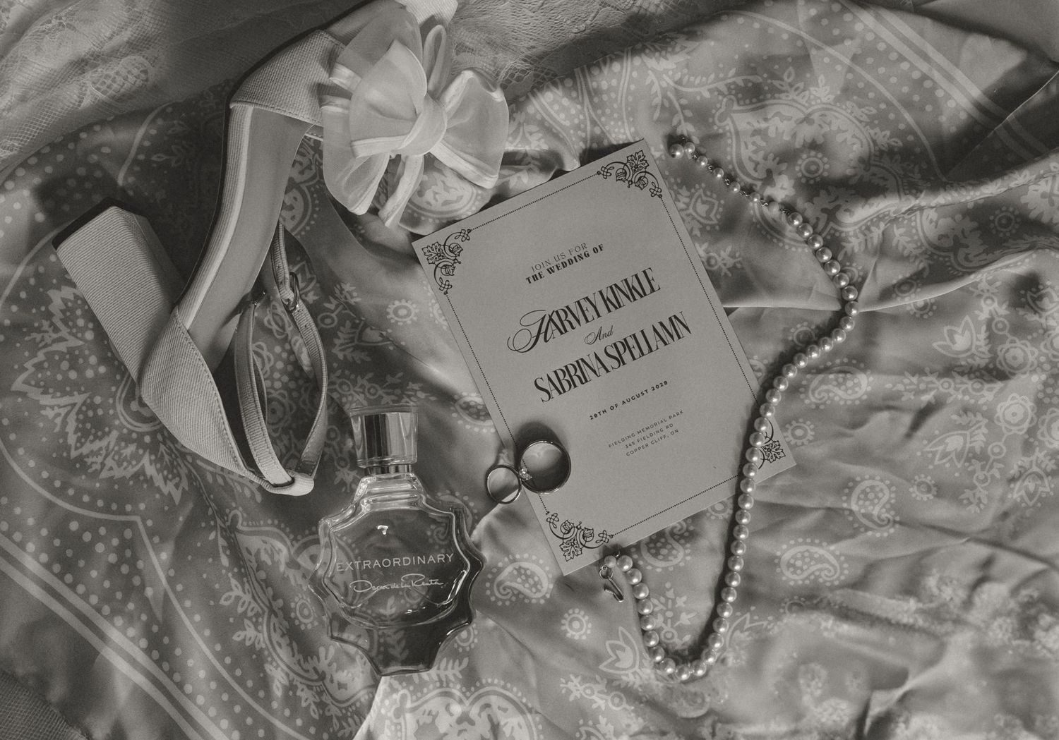 Black and white flat lay of wedding details including perfume, pearls, and ceremony program on lace fabric.