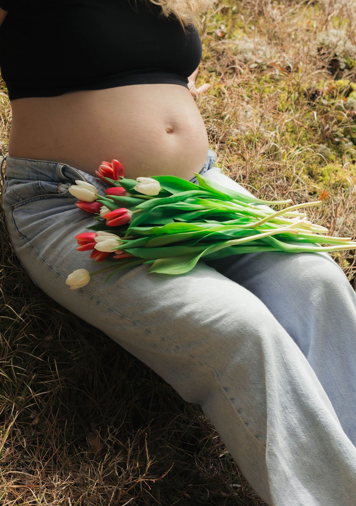 Pregnant woman sitting outdoors in jeans and crop top, holding a bouquet of red and white tulips on her baby bump.