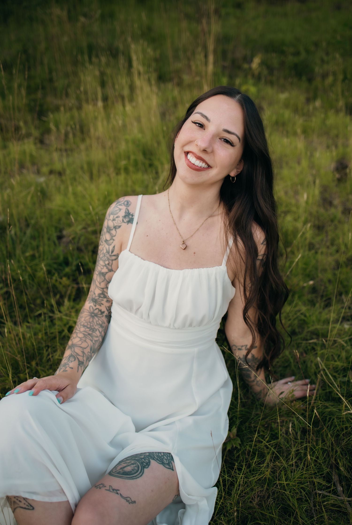 Smiling tattooed woman in white dress sitting in green grass field outdoors.