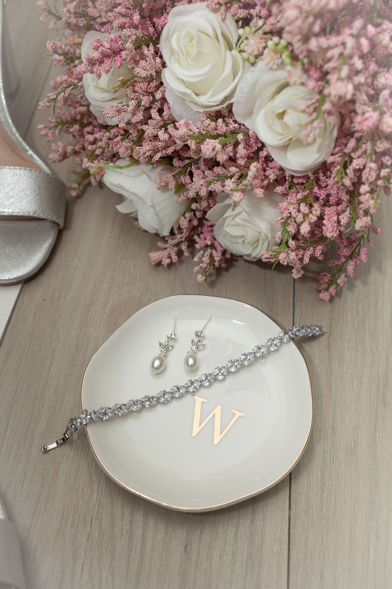 Crystal bracelet and pearl drop earrings on monogram dish beside white roses and pink wildflower bridal bouquet.