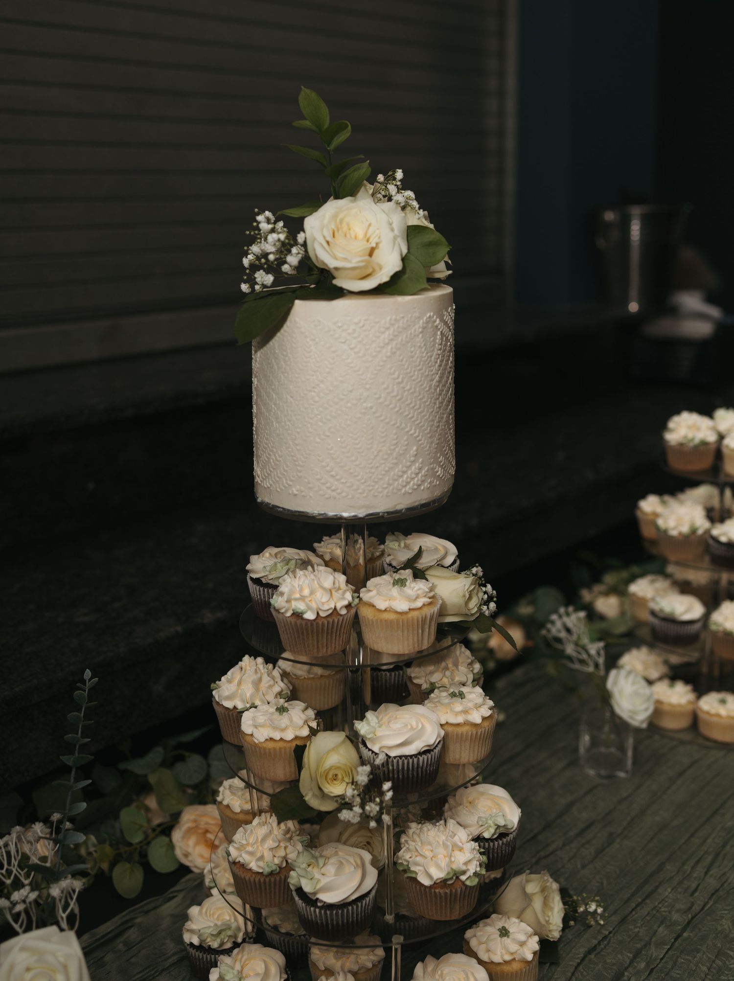 Elegant wedding cake tower with white floral-topped single tier cake above tiered cupcake stand with cream frosting.